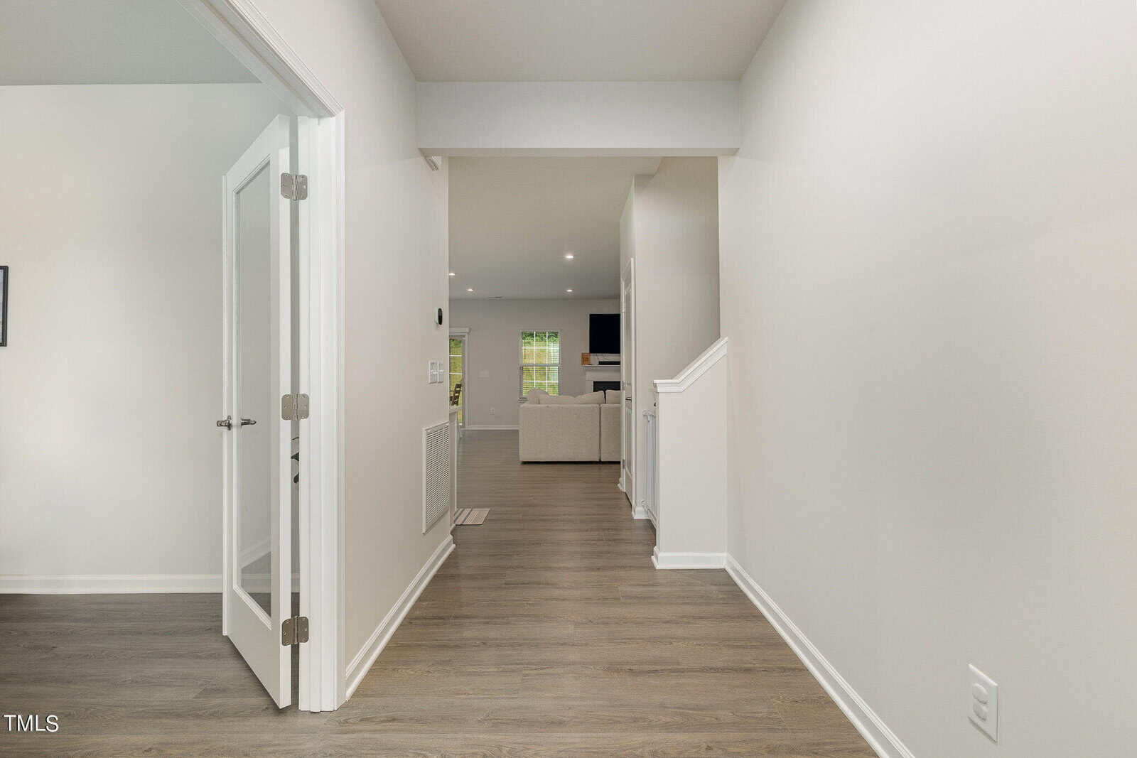 A hallway with light-colored walls and wood flooring leads to a bright living area with a sofa. A room with glass French doors is on the left, and a staircase with a white banister is on the right.