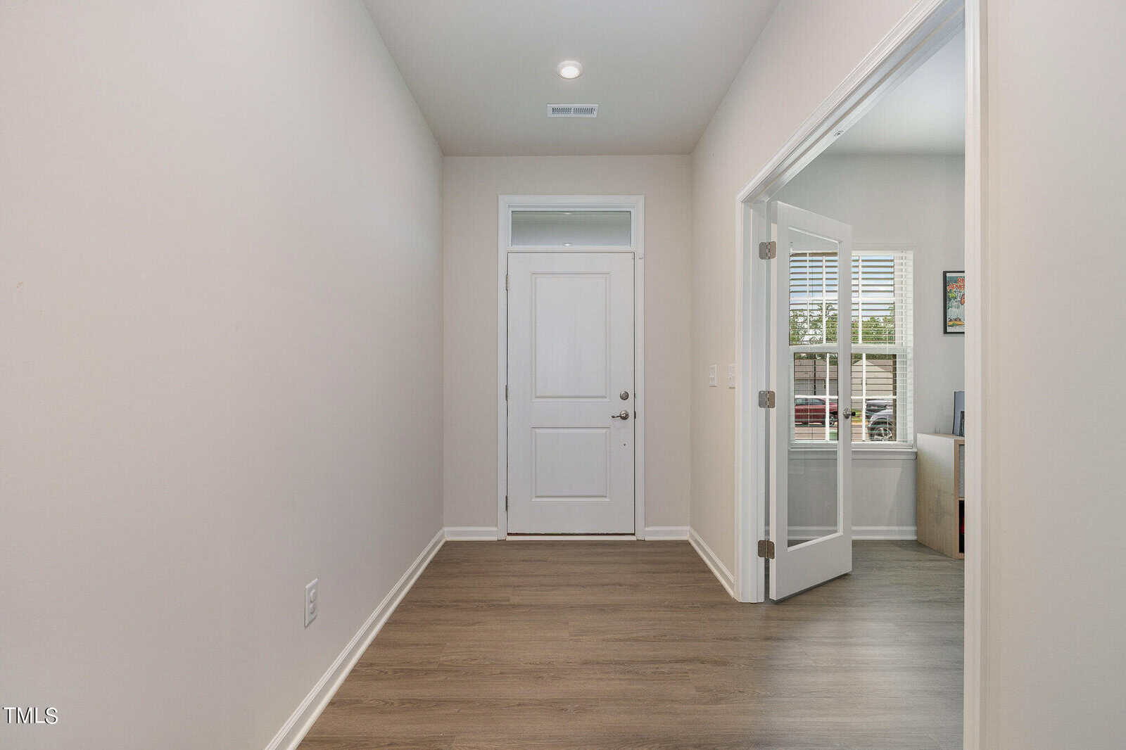 A hallway with light-colored walls and wood flooring leads to a white front door. To the right, glass double doors open to a home office with a window and desk. Ceiling lights brighten the space.