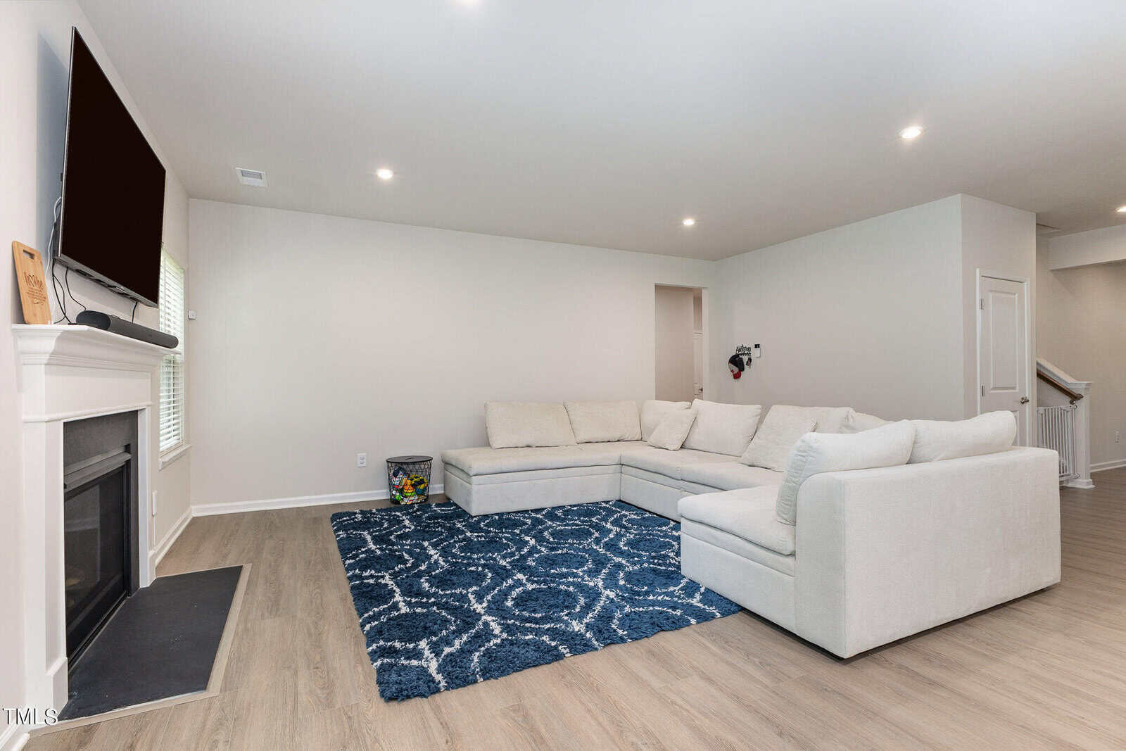 A modern living room with a white sectional sofa, a blue patterned rug, a wall-mounted TV above a fireplace, and light wood flooring. The space has minimal decor and neutral-colored walls.