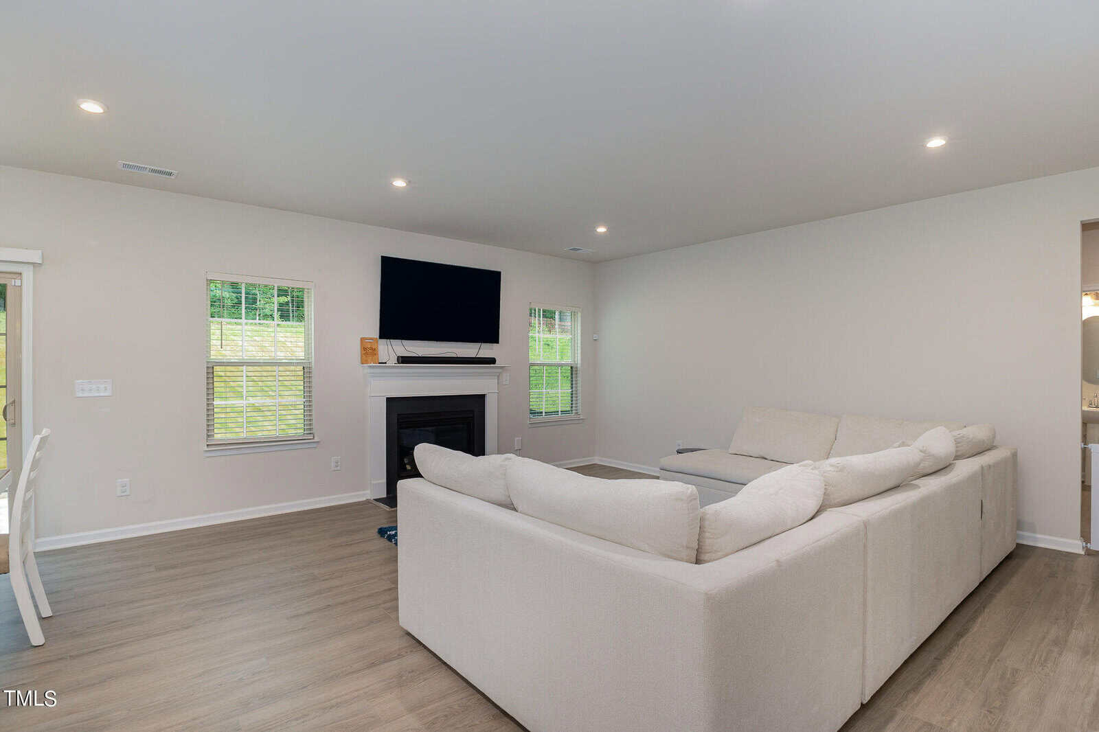 A spacious living room with light wood flooring, a large beige sectional sofa, fireplace with a TV above it, and windows letting in natural light. Walls are painted white and the decor is minimal.