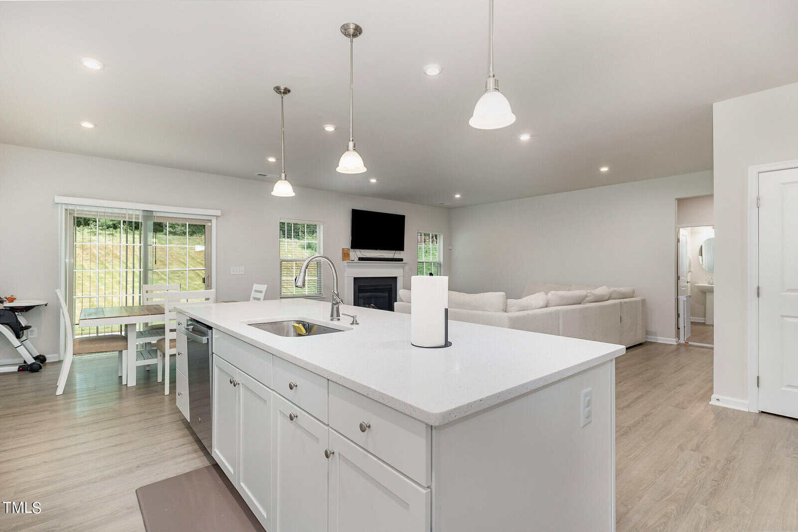 A modern open-concept living space with a white kitchen island, pendant lights, a dining area with sliding glass doors, a cozy living room with a sectional sofa, and a wall-mounted TV above a fireplace.