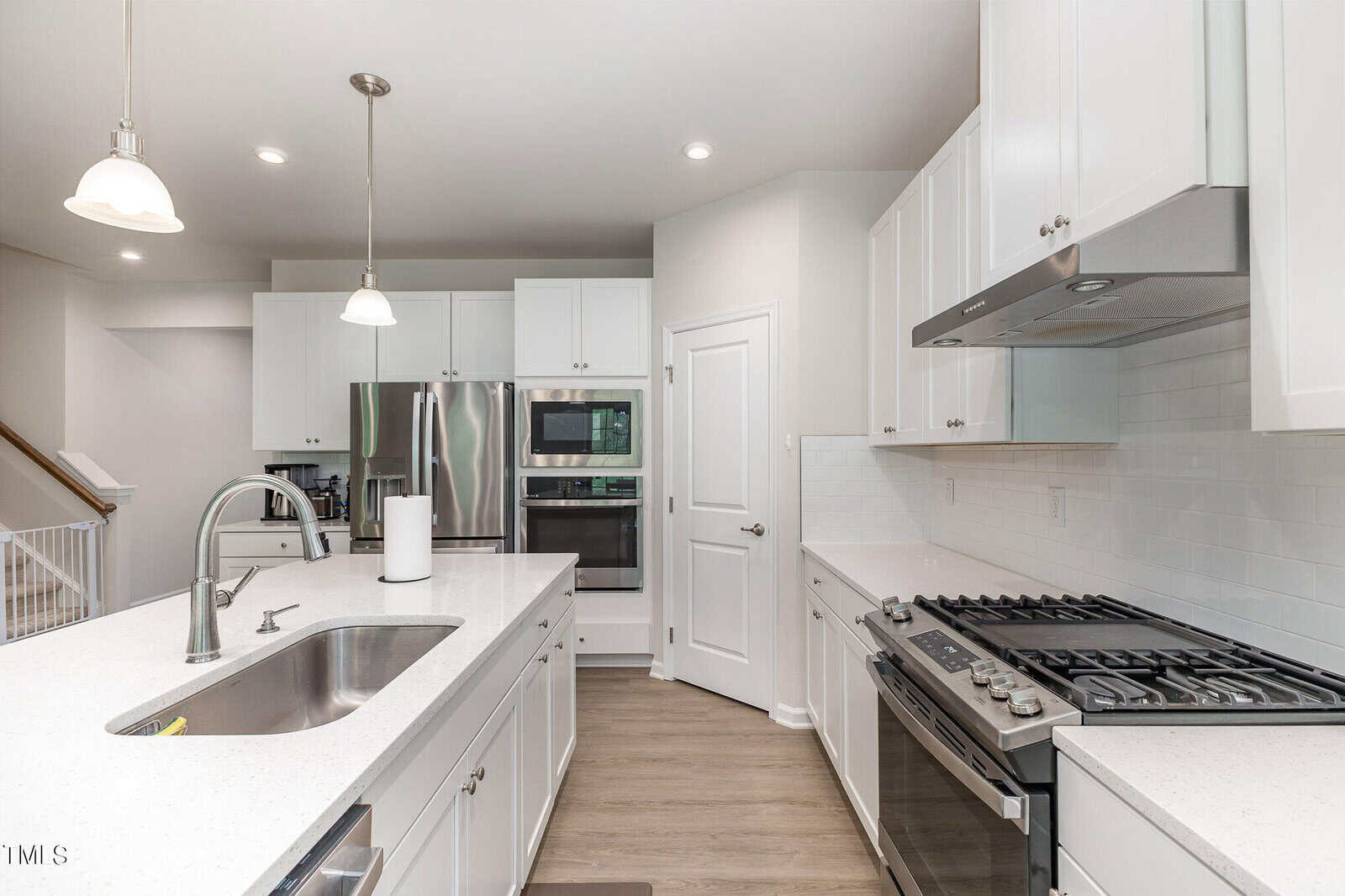 Modern kitchen with white cabinets, stainless steel appliances, a gas stove, double wall ovens, an island with a sink and faucet, pendant lights, and light wood flooring. A staircase is visible to the left.