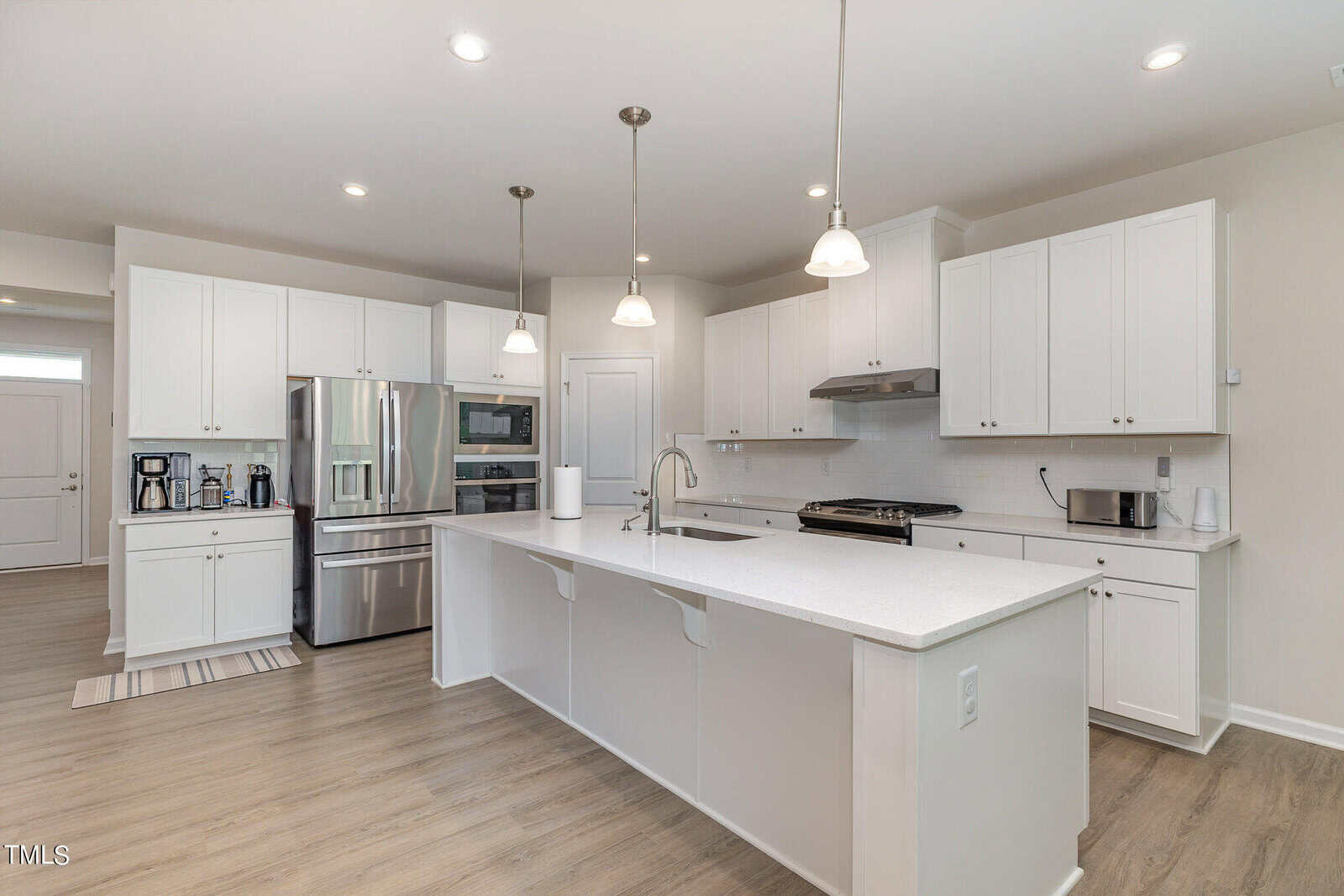 Modern kitchen with white cabinets, stainless steel appliances, a large island with a sink, pendant lighting, light wood flooring, and various small appliances on the counters.