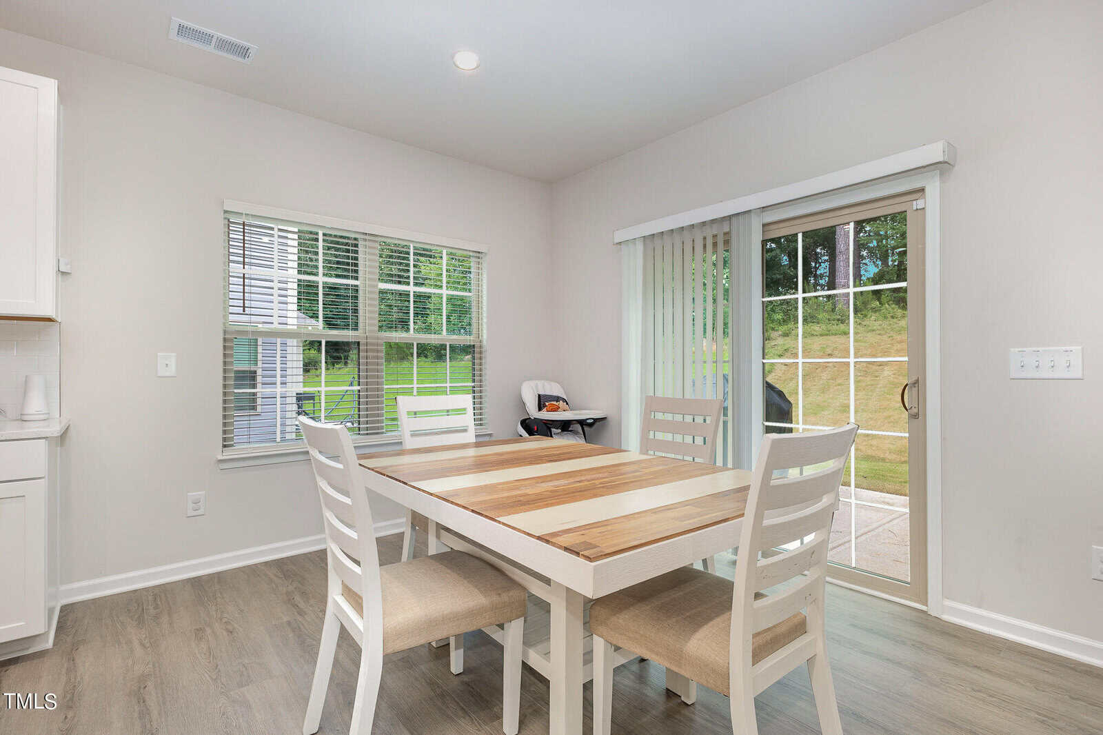 Bright dining area with a wooden table and four chairs, large window, sliding glass door to a patio, and high chair in the corner. Light wood floors and pale walls create a clean, airy atmosphere.