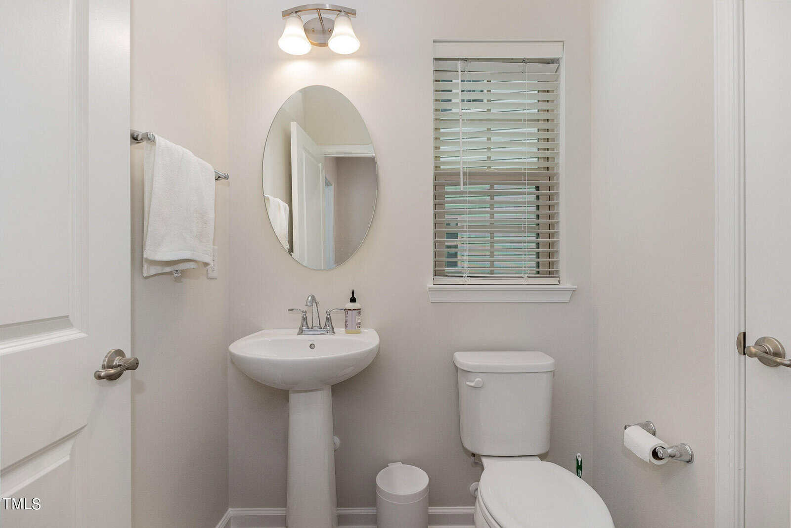 Small bathroom with white walls, pedestal sink, oval mirror, towel on a rack, toilet with a closed lid, toilet paper holder, trash can, and window with blinds. Overhead light above the mirror.