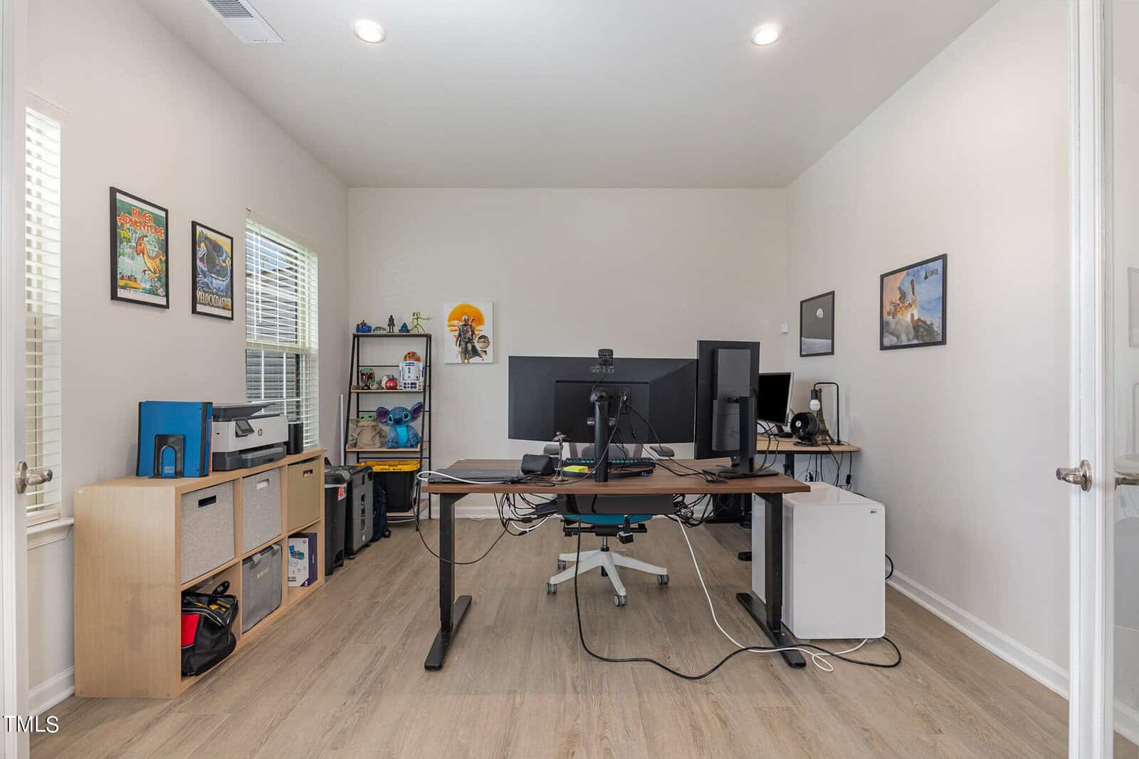 Home office with dual monitors, various electronics, shelves with bins, toys, books, and art on the walls. Light wooden floor and two windows letting in natural light.