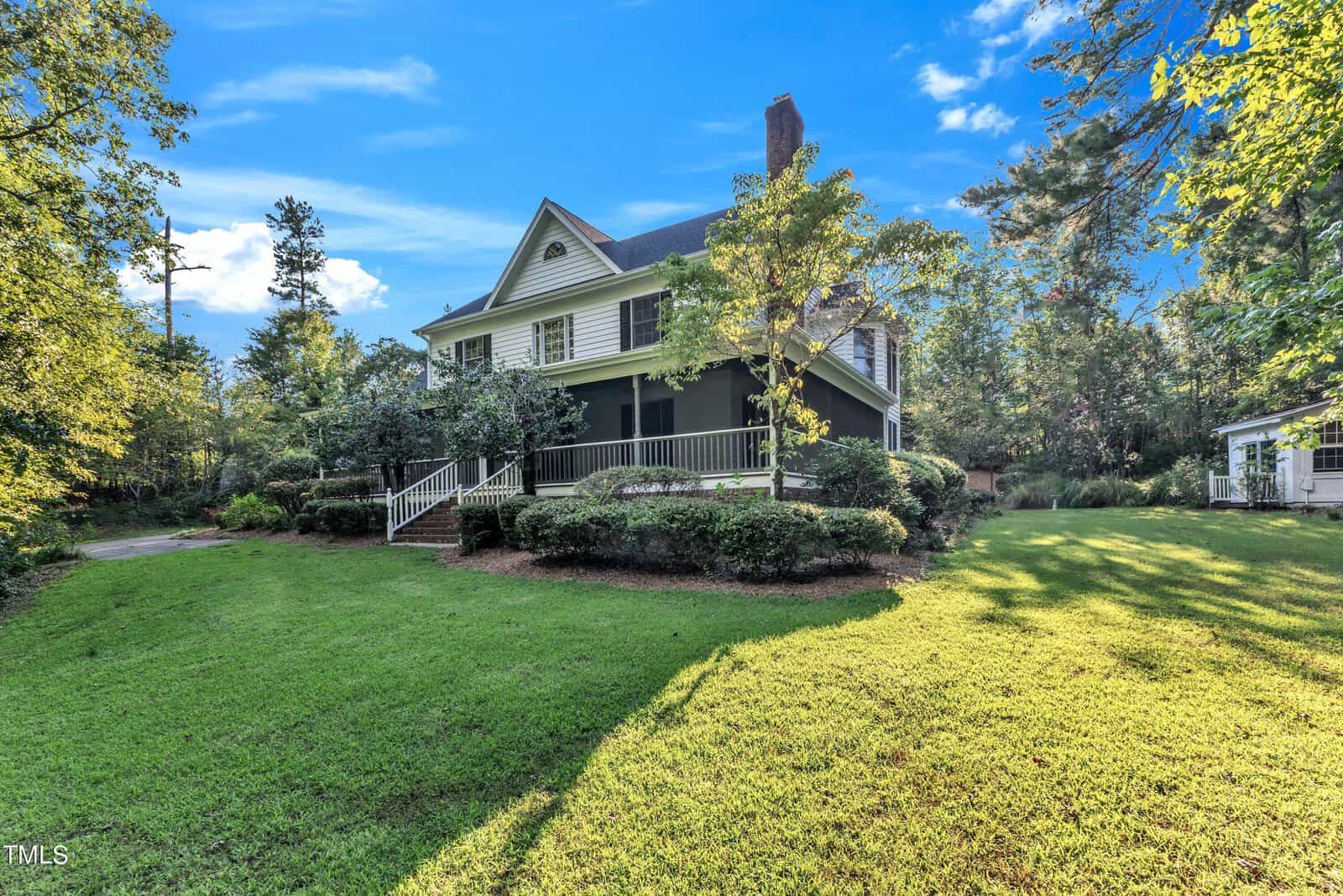 A picturesque two-story house with a wraparound porch surrounded by lush greenery and trees under a bright blue sky. The well-maintained lawn extends to the front and side of the house. The house exterior is dark with white trim, and the scene is bathed in sunlight.