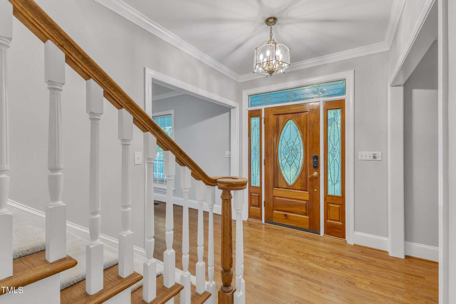 A brightly lit foyer with a wooden staircase on the left, leading up to the second floor. The front door features stained glass windows with intricate designs, and a chandelier hangs from the ceiling. White walls and a hardwood floor complete the elegant look.