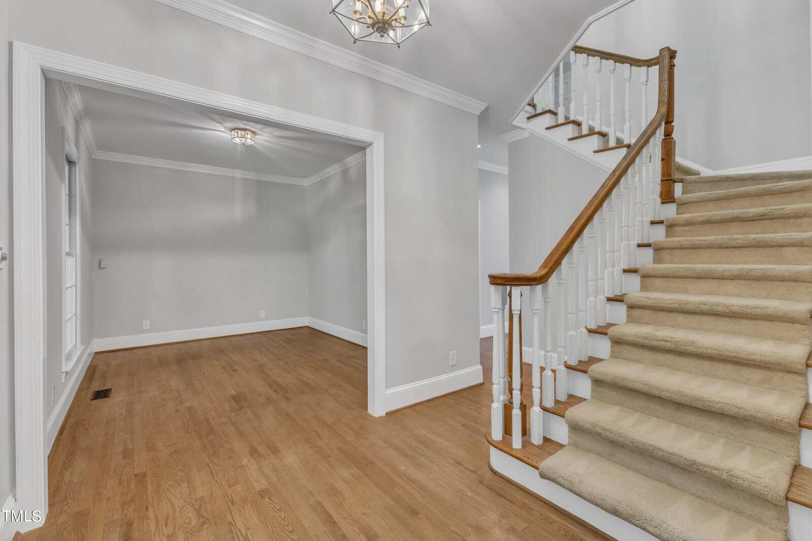 A spacious interior featuring a carpeted staircase with a wooden handrail on the right and a hardwood-floored room with large windows on the left. The walls are painted light gray, and both rooms are illuminated by elegant ceiling light fixtures.