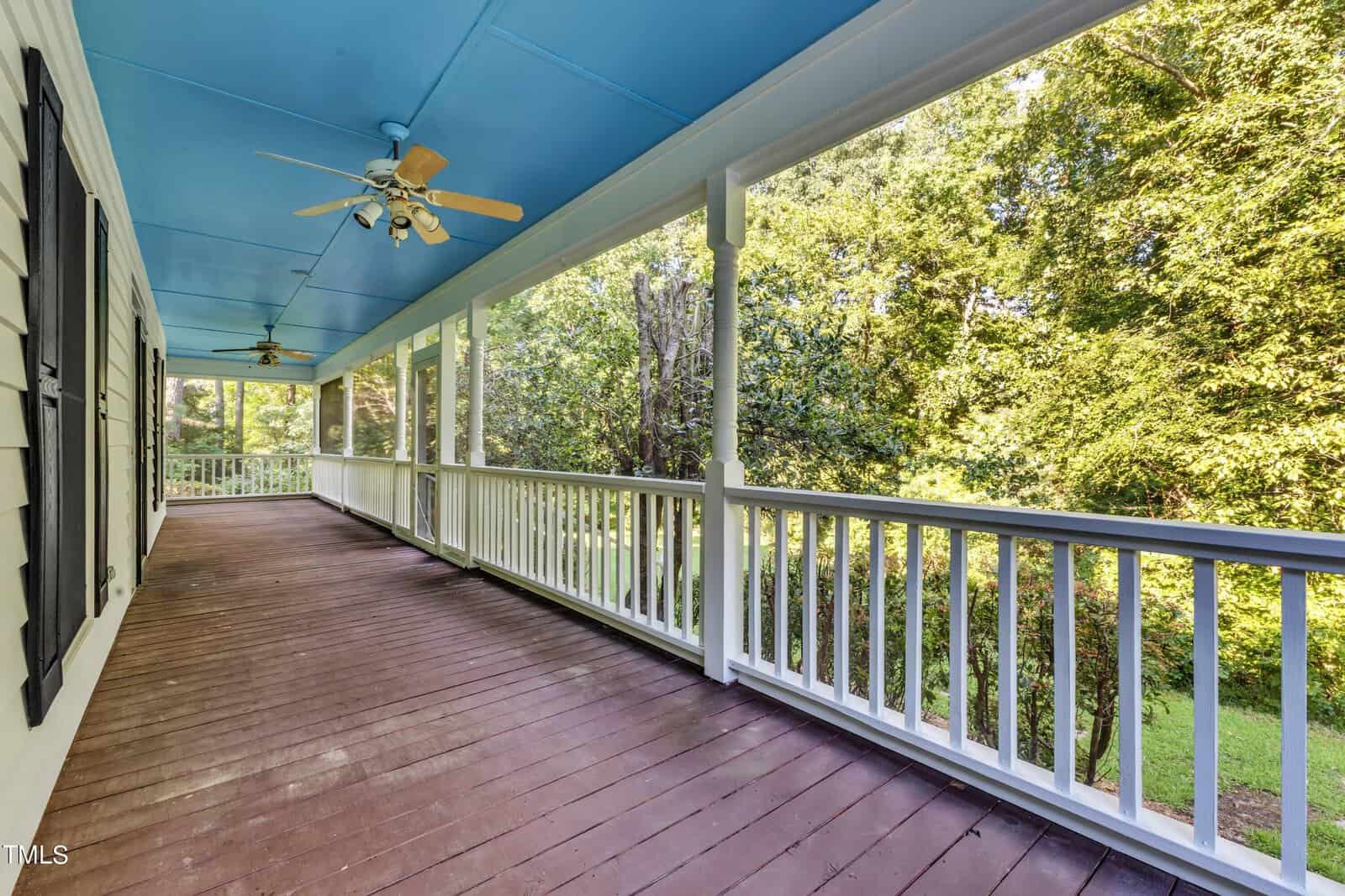 A spacious porch with a blue ceiling and wooden flooring, bordered by a white railing. Two ceiling fans with lights hang from above. The porch offers a view of lush green trees and a grassy yard on the right side.