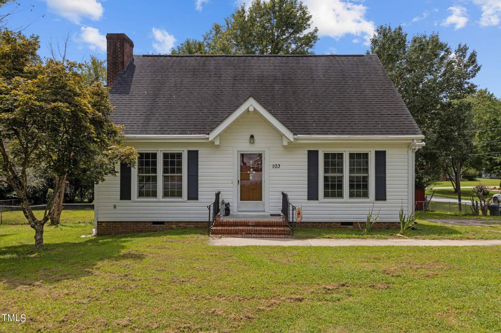 A charming white house with a steep gray roof and brick chimney, featuring a front porch with steps, large windows, and a well-kept lawn with a tree. Surrounded by greenery under a partly cloudy blue sky.