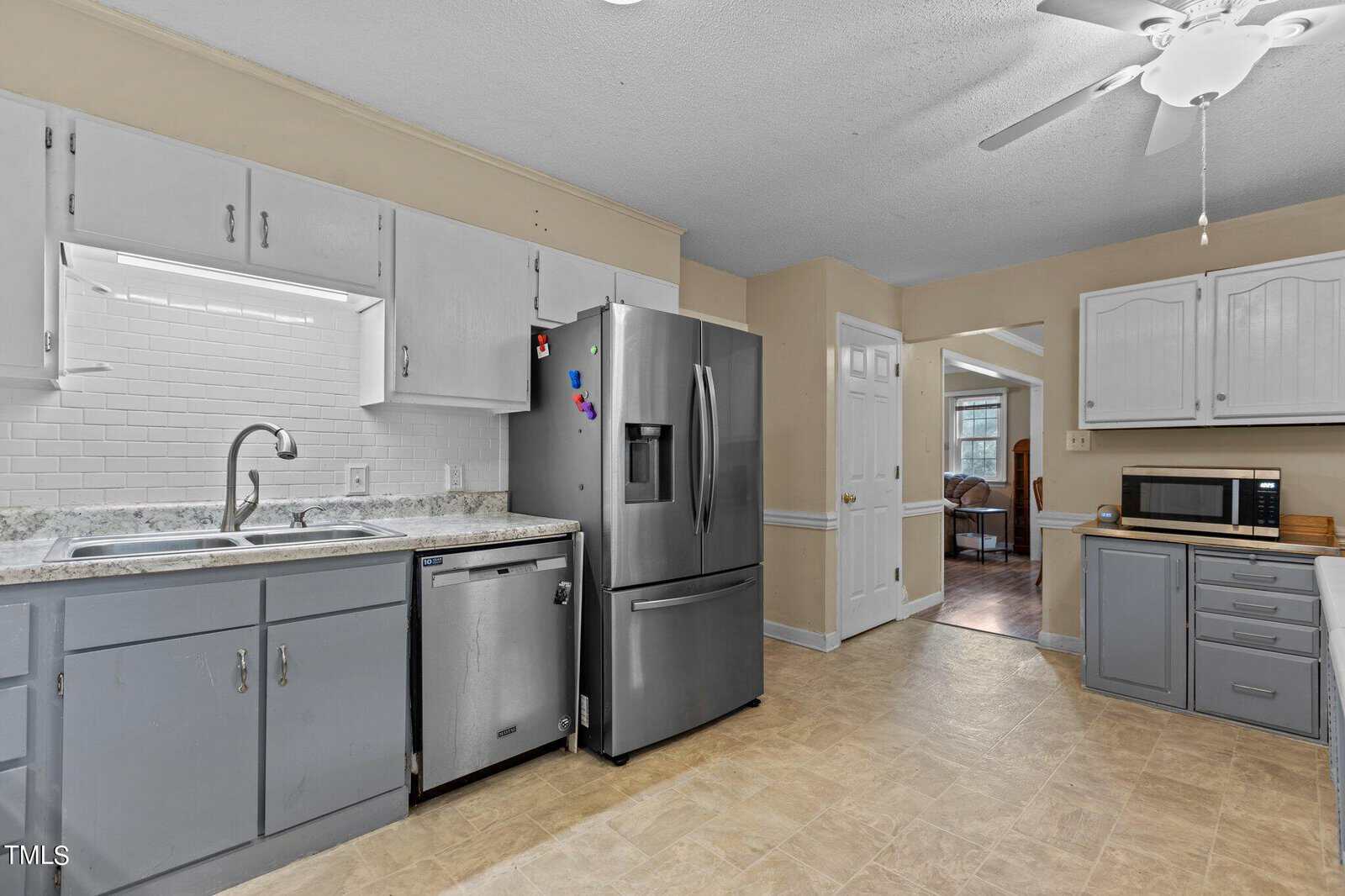 A kitchen with a mix of white and gray cabinets, a stainless steel refrigerator, and a dishwasher. There is a sink under a window with white subway tile backsplash. A microwave sits on the counter next to a doorway leading to another room.