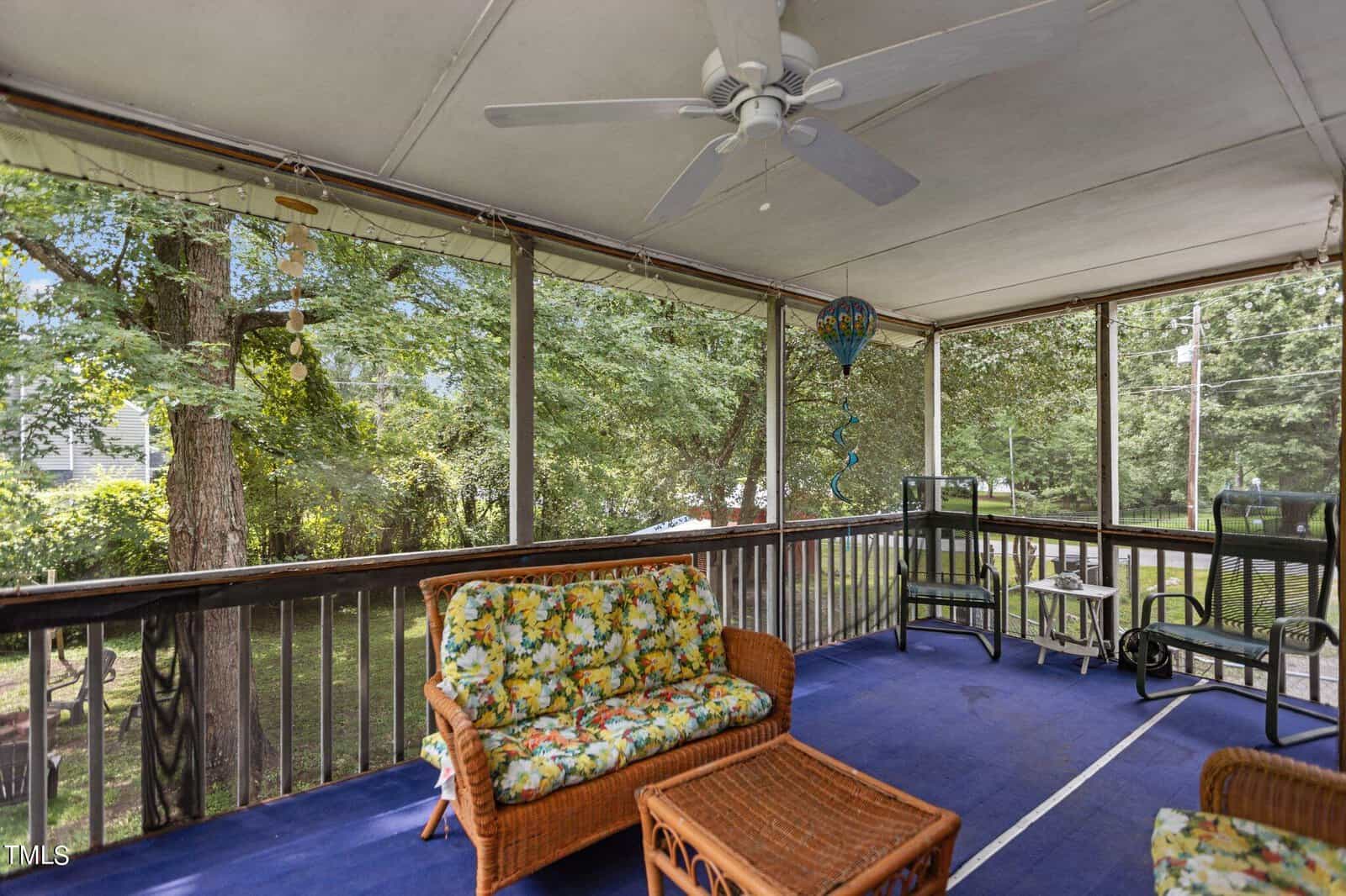 A cozy screened porch with a ceiling fan, adorned with wicker furniture featuring floral cushions. A blue carpet covers the floor, and trees surround the area outside the screened windows. A heart-shaped wind chime hangs from the ceiling.