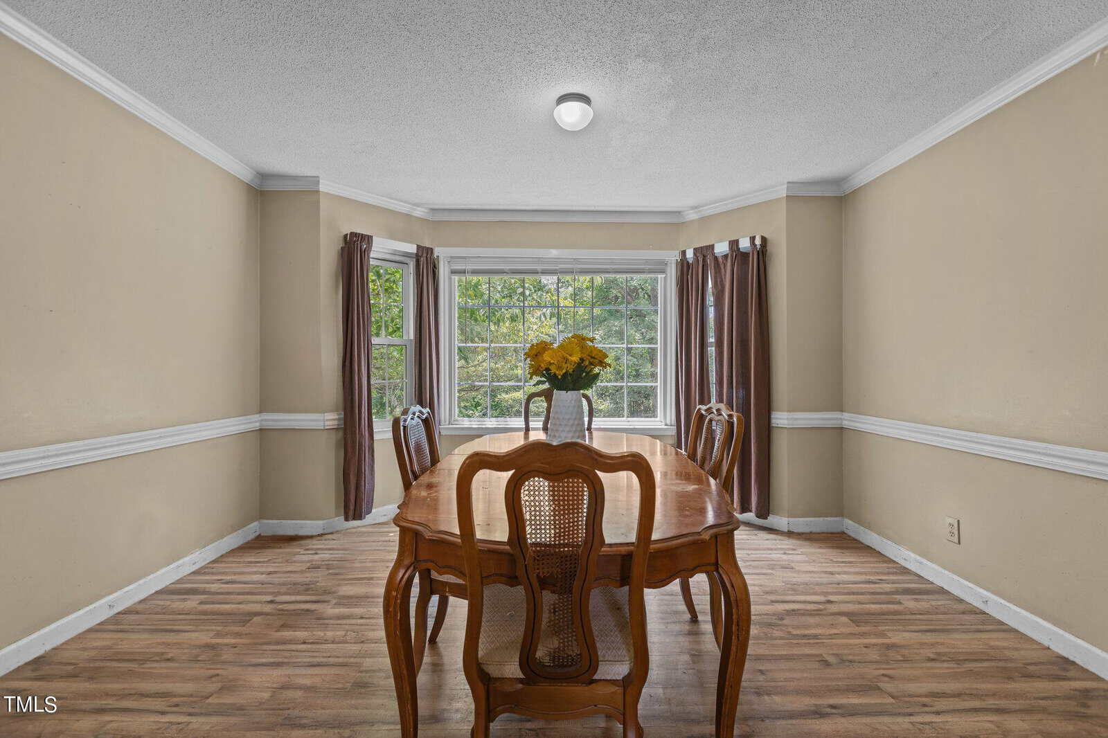 A dining room with a wooden table and four chairs. A vase of yellow flowers is centered on the table. The room has beige walls, a large bay window with brown curtains, and wooden flooring. Natural light fills the space.