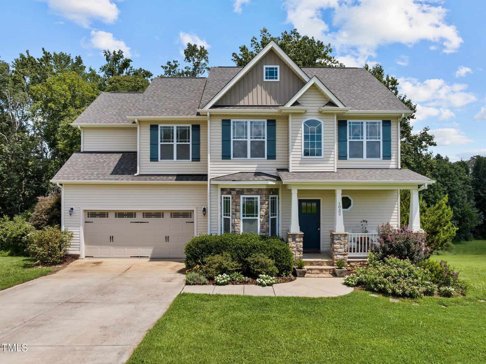 At 1032 Snapdragon Drive in Wake Forest, a two-story house with a gray roof and beige siding stands amidst a green lawn. It features a two-car garage, a small front porch with columns, and landscaped bushes and trees. The sky is partly cloudy.