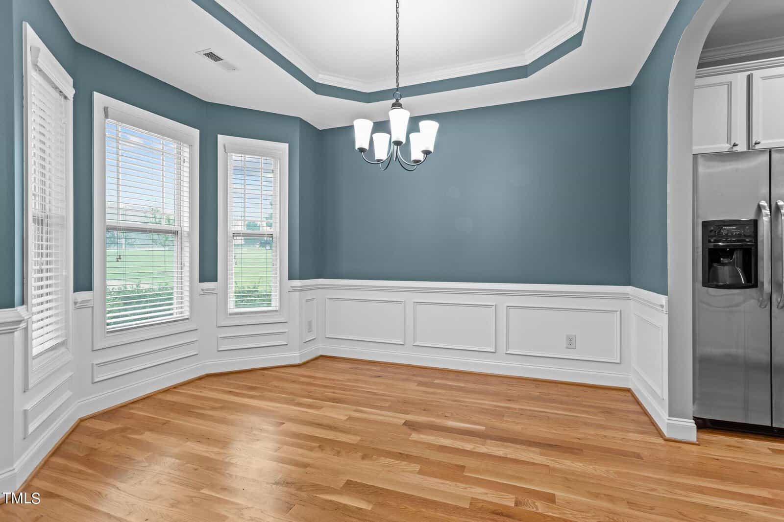 A bright dining area with green walls, white wainscoting, and a wooden floor. Three large windows allow natural light to enter. A chandelier hangs from a recessed ceiling. A stainless steel refrigerator is visible in the background.