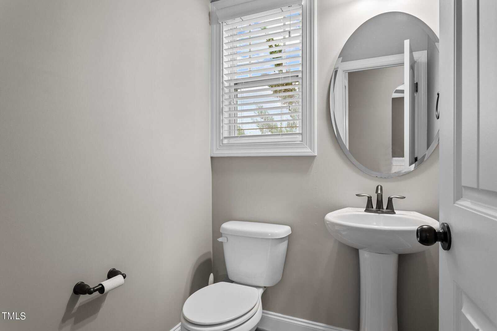 A small, neatly arranged bathroom with light gray walls. It features a white toilet, an oval mirror above a pedestal sink, and a window with horizontal blinds letting in natural light. A toilet paper holder is mounted on the wall.