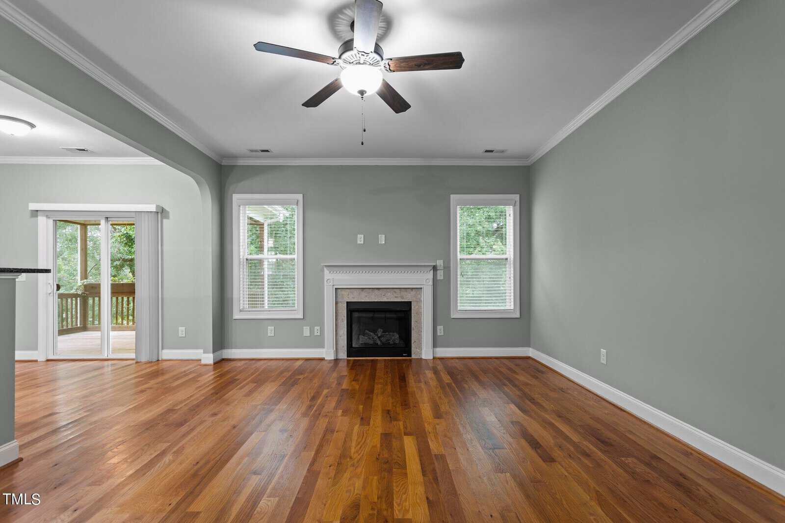 A spacious living room with light green walls, hardwood flooring, and a white fireplace centered between two windows. A ceiling fan with lights is above, and a doorway leads to a wooden deck outside.