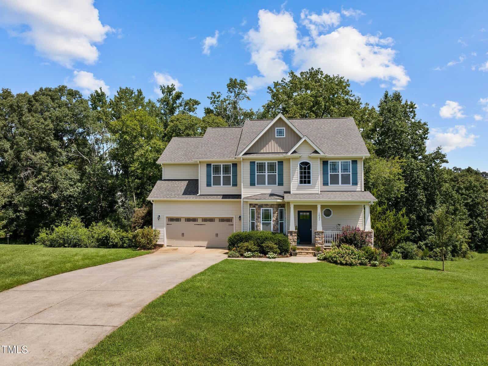 A two-story suburban house with a gray roof and beige siding sits at the end of a concrete driveway. It is surrounded by a well-manicured lawn and trees. The sky is blue with scattered clouds.