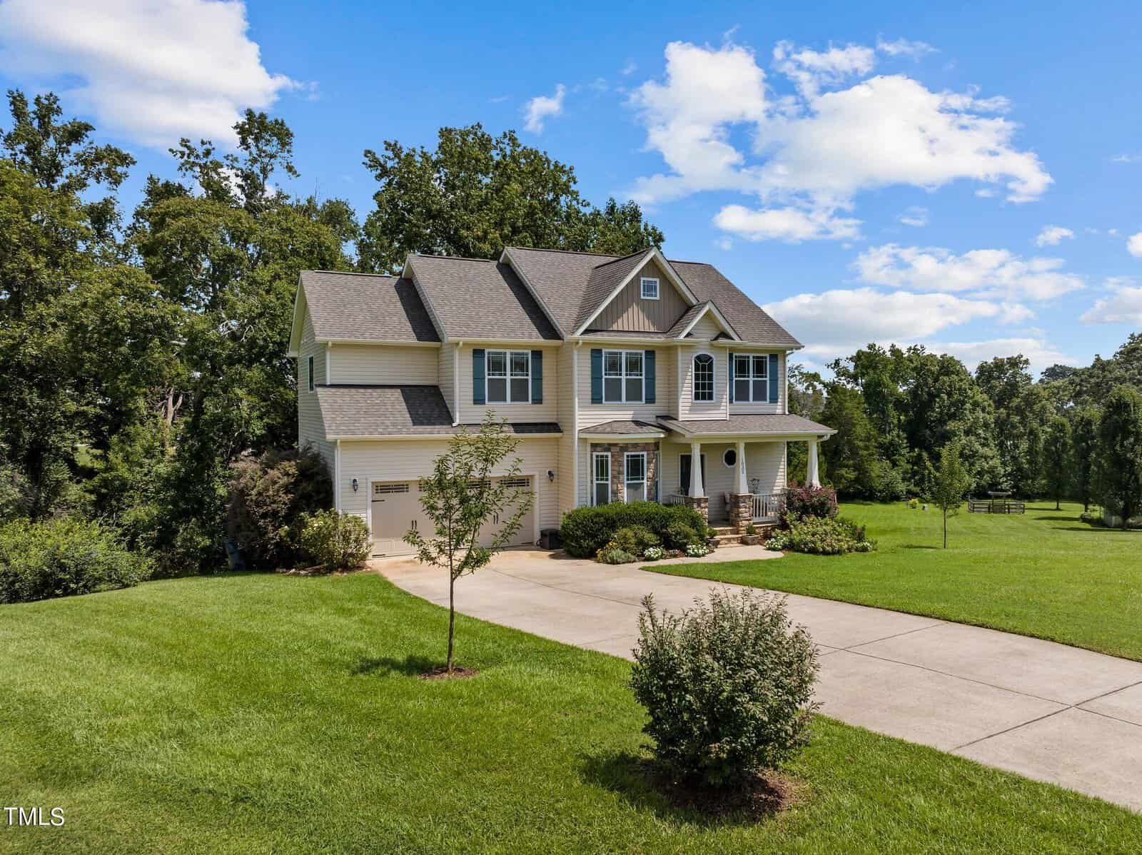 A two-story house with a gray roof and light-colored siding sits surrounded by a lush green lawn. The driveway leads up to a garage on the left. The sky is clear, with a few clouds and trees in the background.
