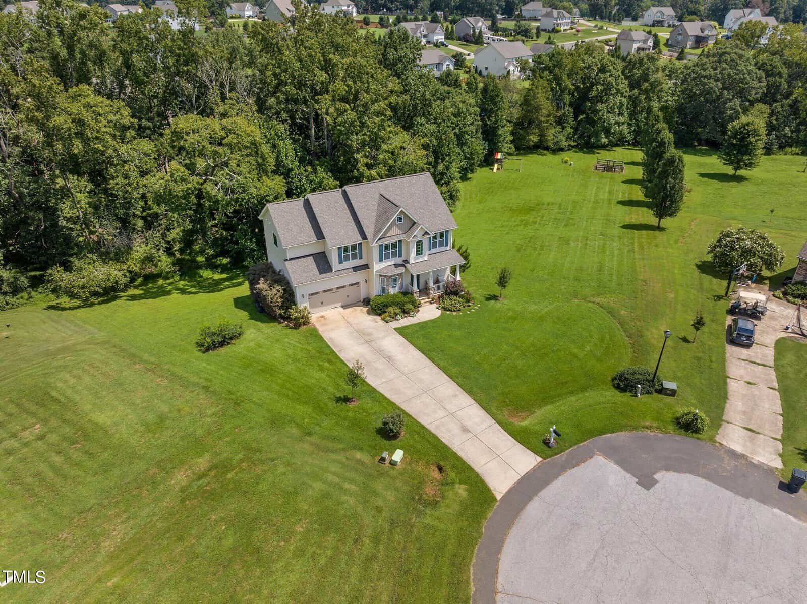 Aerial view of a large two-story house with a spacious lawn, situated at the end of a cul-de-sac. The property is surrounded by trees, and there are several other houses visible in the background.