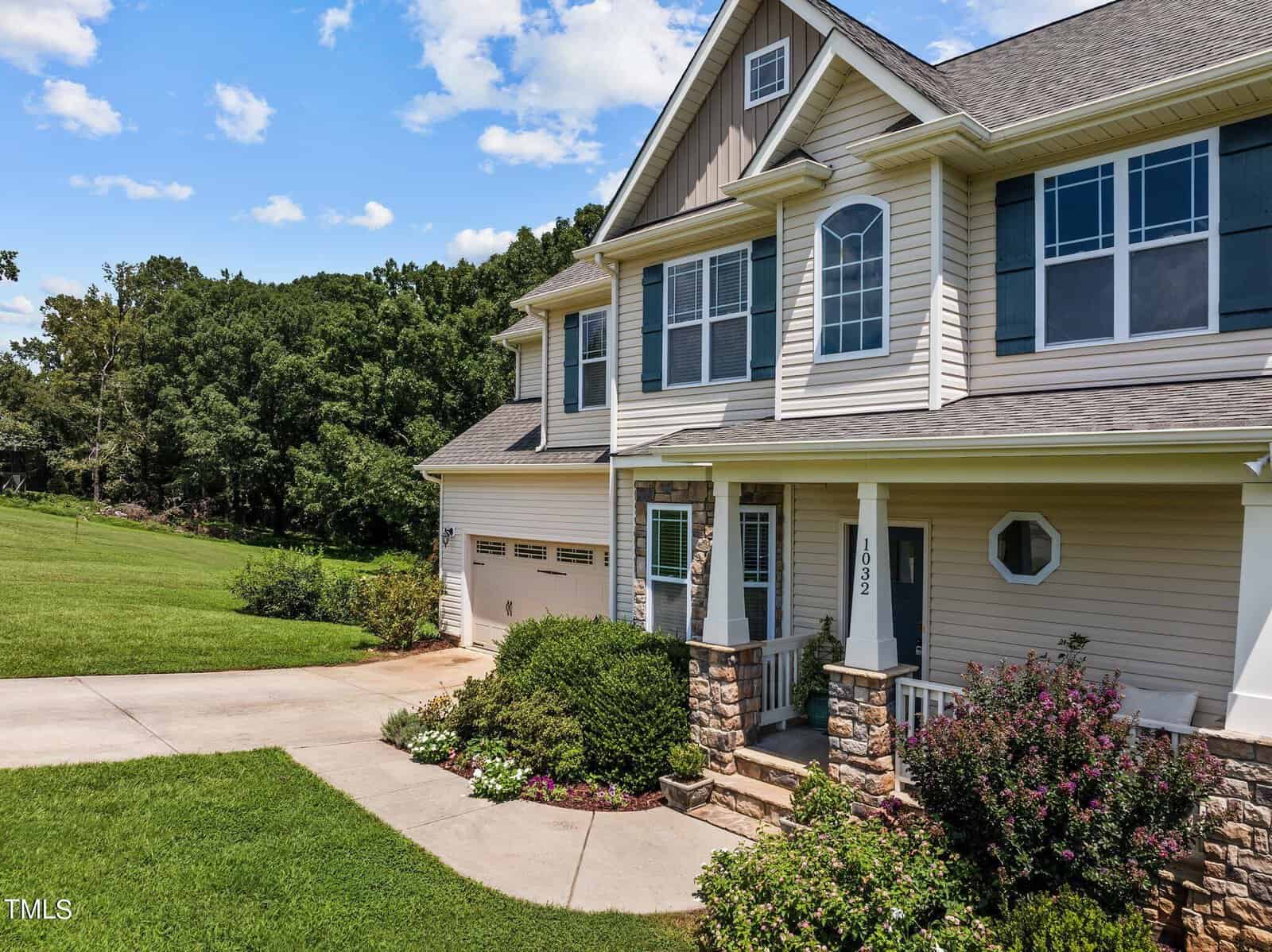 Two-story house with stone accents and blue siding. It has a two-car garage, front porch with columns, and a well-maintained lawn with bushes. The driveway curves to the left, and trees are visible in the background under a partly cloudy sky.