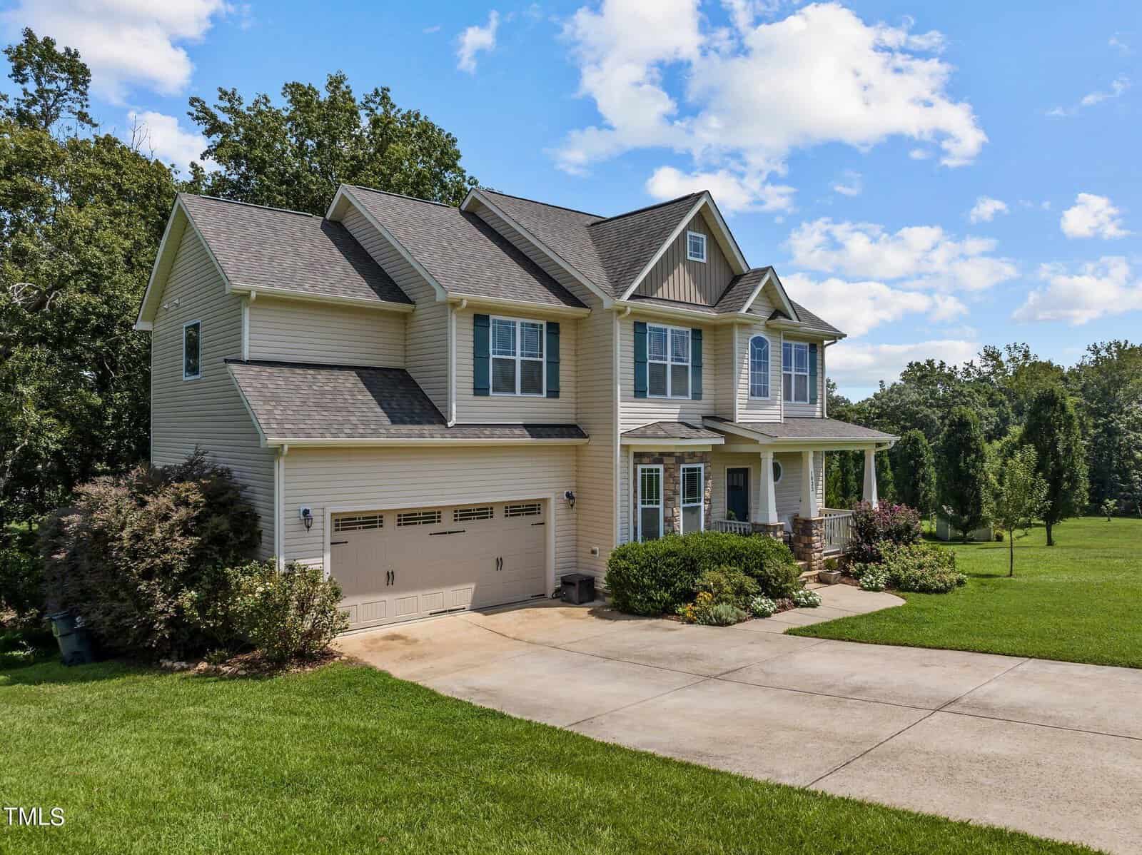 A two-story beige house with a large driveway and attached garage, surrounded by a well-maintained green lawn and mature trees under a clear blue sky with scattered clouds.