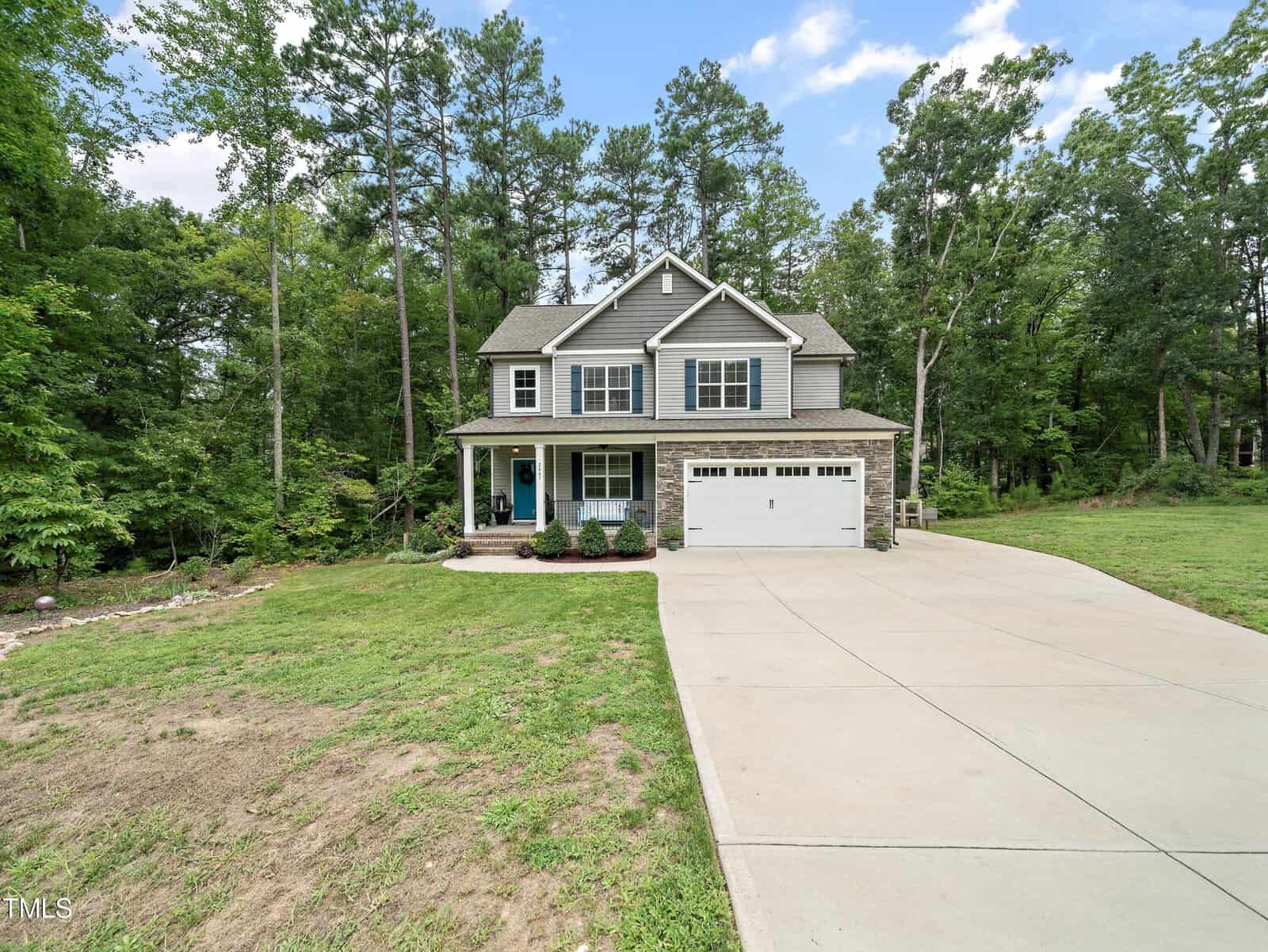 Nestled among tall green trees, the two-story gray house at 2467 Golden Forest Drive in Franklinton boasts a welcoming porch. It features a two-car garage and a wide concrete driveway, while the lawn, partially grass-covered, extends to the edge of a wooded area under a blue sky.