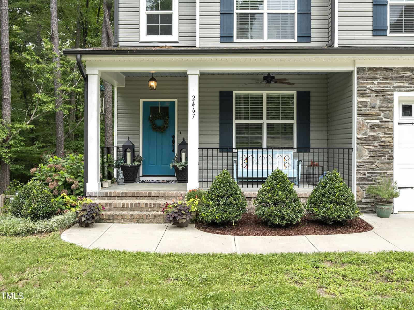 A house with gray siding and stone accents features a covered porch. The porch has a blue door with a wreath, two black lanterns, and a cushioned bench. Neatly trimmed shrubs line the walkway in the front yard.
