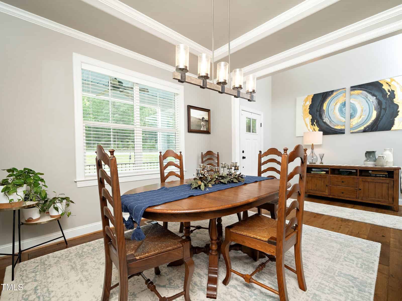 A dining room with a wooden table and six chairs, adorned with a navy blue table runner and centerpiece. A modern chandelier hangs above. The room features a large window with blinds, artwork on the wall, and a sideboard with decor items.
