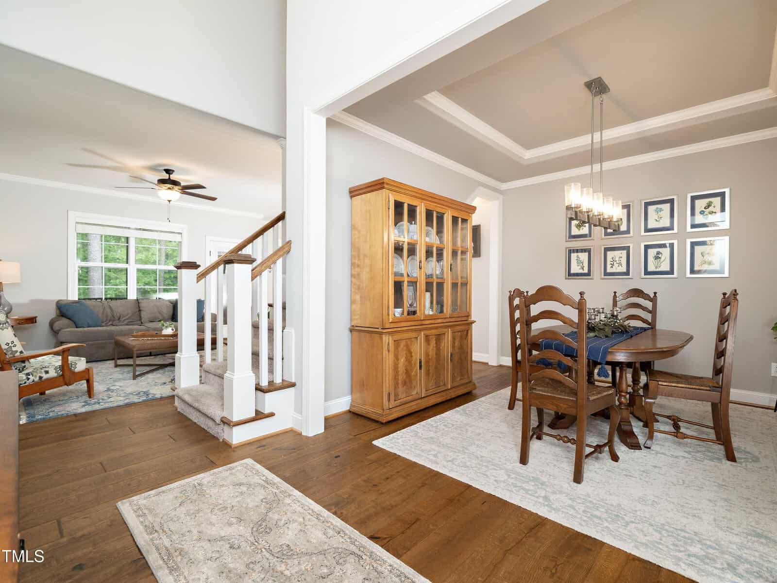 A bright, open-concept living and dining area with hardwood floors. The dining room features a wooden table with chairs and a matching cabinet. The living room has a gray sofa, armchair, and a ceiling fan. Artwork adorns the walls.