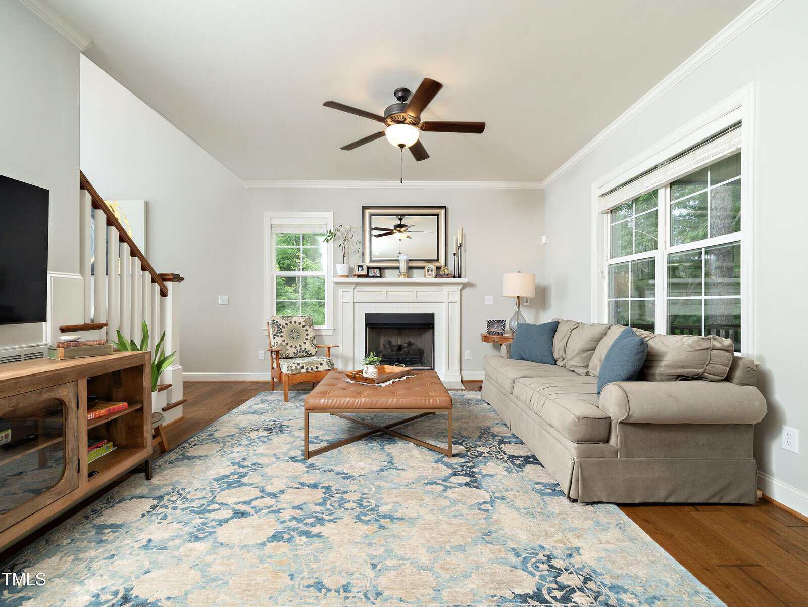 A modern living room with a gray sectional sofa, blue patterned rug, and wooden coffee table. A fireplace with a mirror above it is centered on the wall. Ceiling fan and large windows provide light. A staircase is visible to the left.