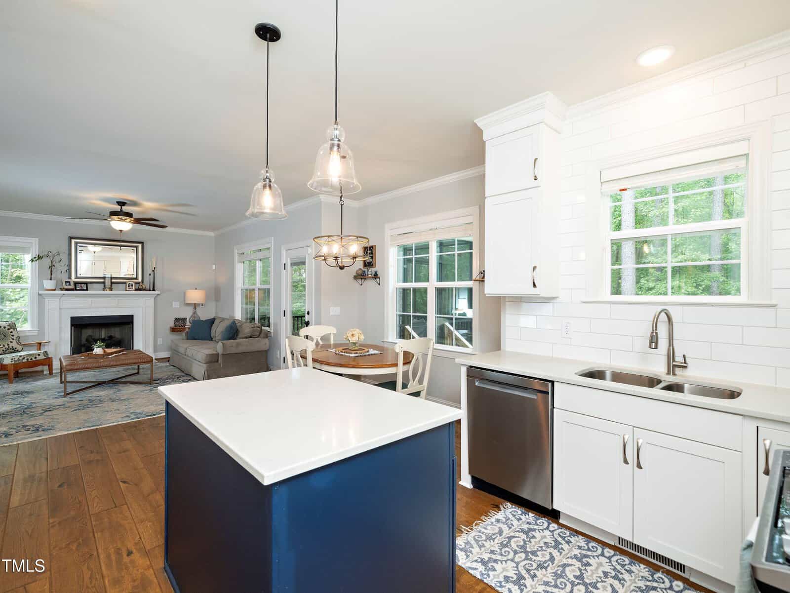 Modern kitchen and open living area with white cabinets, blue island, pendant lights, stainless steel appliances, and hardwood floors. A dining table sits by large windows, and a cozy sofa faces a fireplace in the adjacent room.