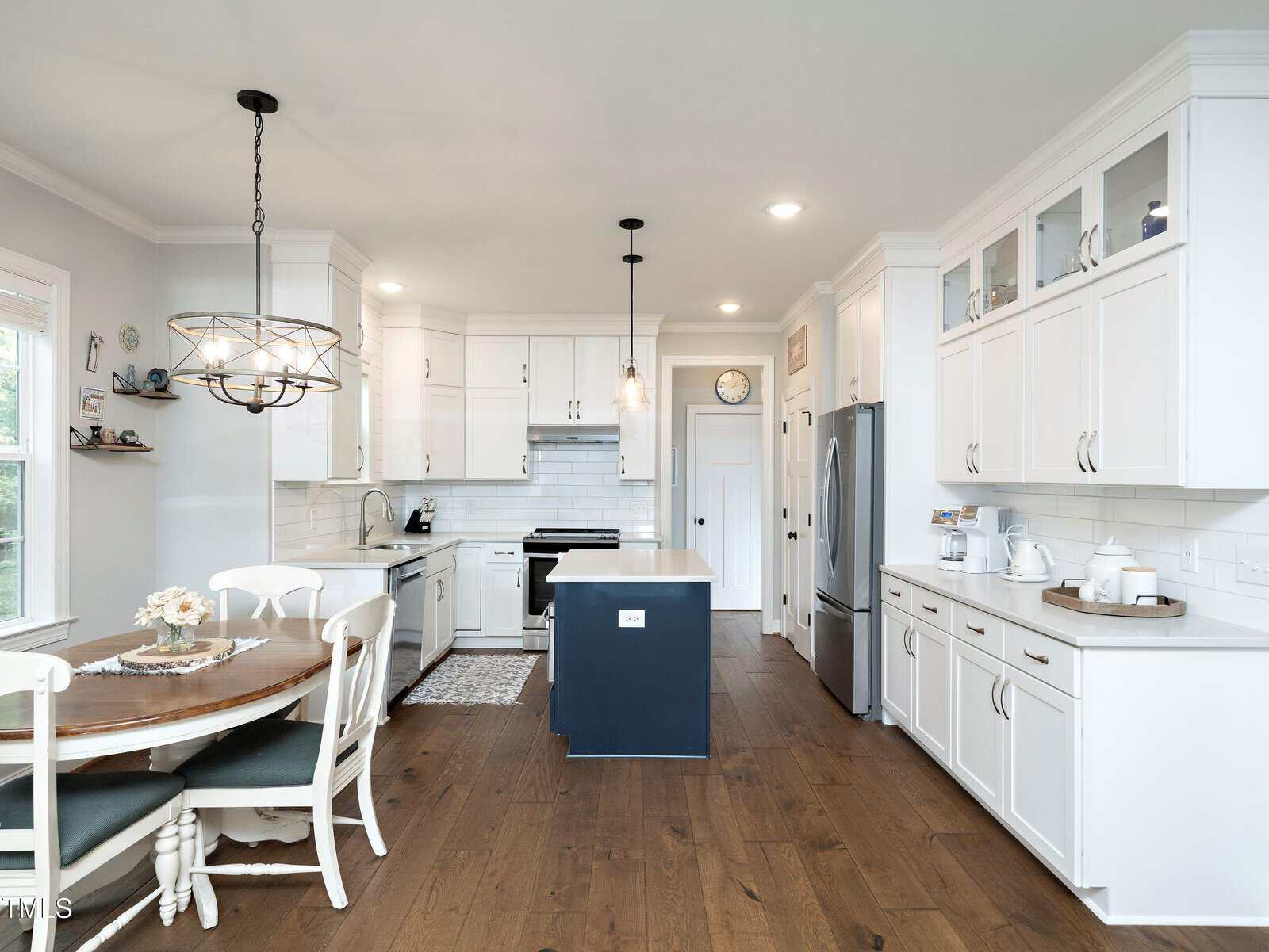 A modern kitchen with white cabinetry, a central island with a dark blue base, stainless steel appliances, and wooden flooring. The dining area features a round wooden table with chairs, under a decorative light fixture.