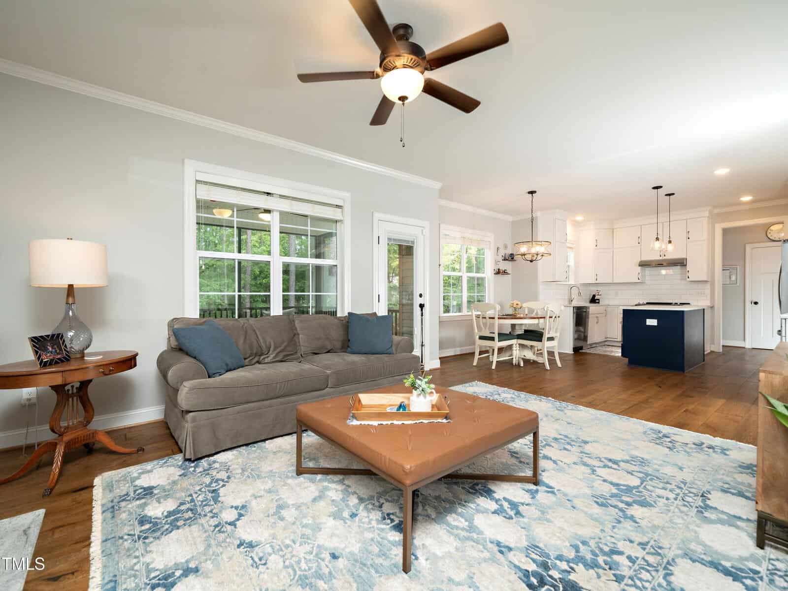 A cozy living room with a gray sofa, wooden coffee table on a blue patterned rug, and a side table with a lamp. In the background, there's a dining area with a round table and a modern kitchen with white cabinets and an island. A ceiling fan is above.