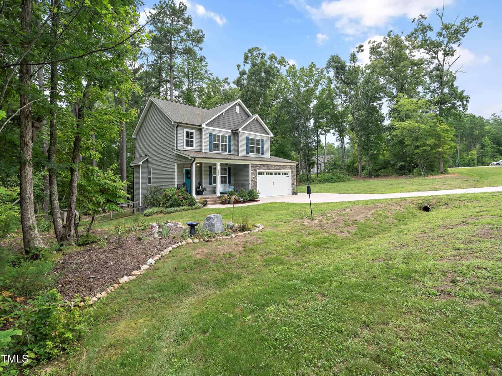 A two-story green house with white trim is surrounded by a lush lawn and trees. It features a front porch, a two-car garage, and a paved driveway. The landscape includes a small garden with rocks and a birdbath.