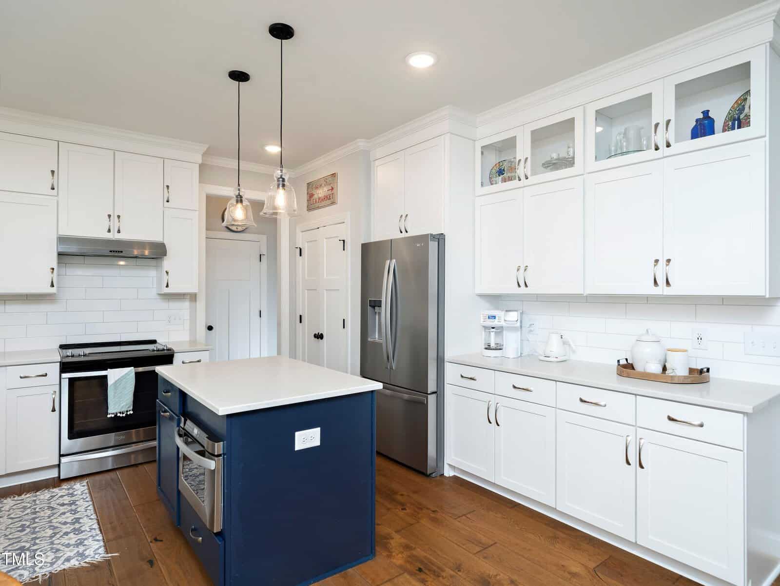 A modern kitchen with white cabinets, a stainless steel refrigerator, and a dark blue island with a white countertop. Pendant lights hang above the island, and a patterned rug lies on the wooden floor.