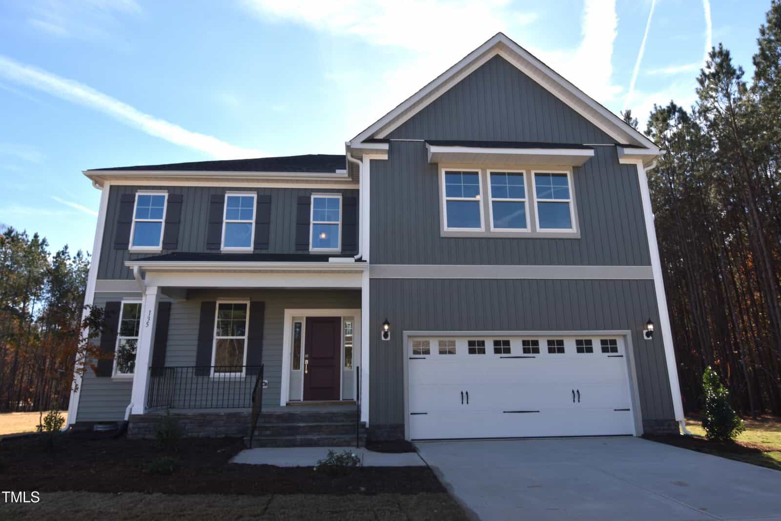 A two-story modern house with gray siding, white trim, a covered front porch, and a double garage. The home is set against a backdrop of tall trees under a clear blue sky.