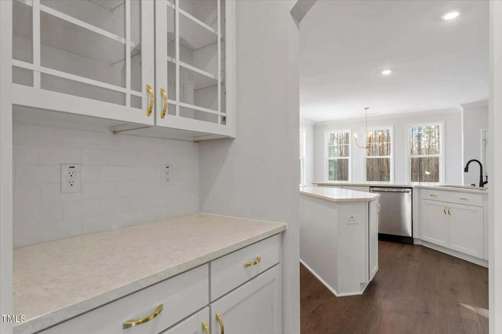 Bright modern kitchen with white cabinets, gold handles, glass-front upper cupboards, light countertops, stainless steel appliances, wood floors, and large windows letting in natural light.