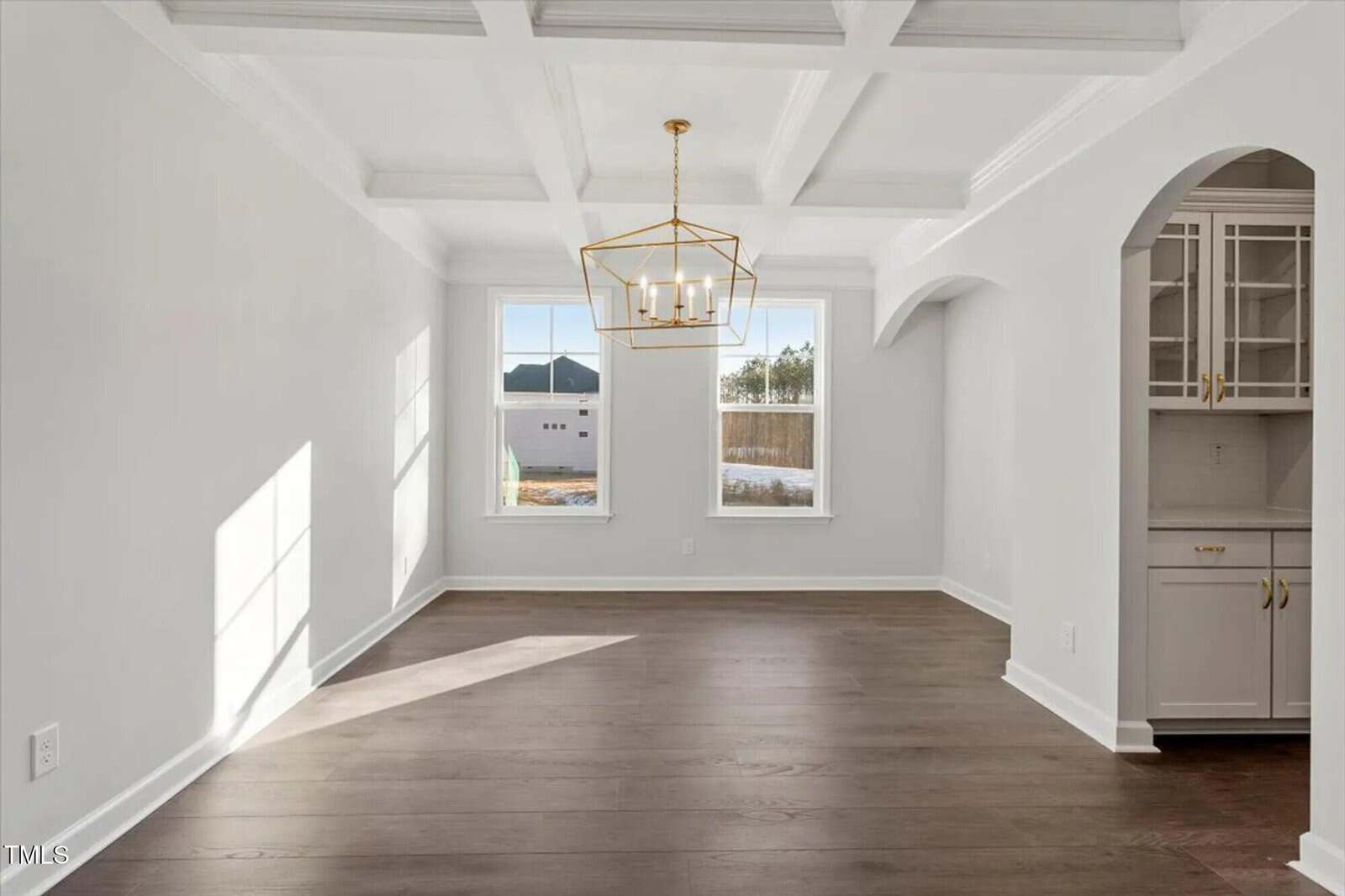 A bright, empty dining room with large windows, dark wood floors, white walls, a coffered ceiling, and a modern gold chandelier. Arched entry leads to a kitchen area with white cabinets and gold hardware.