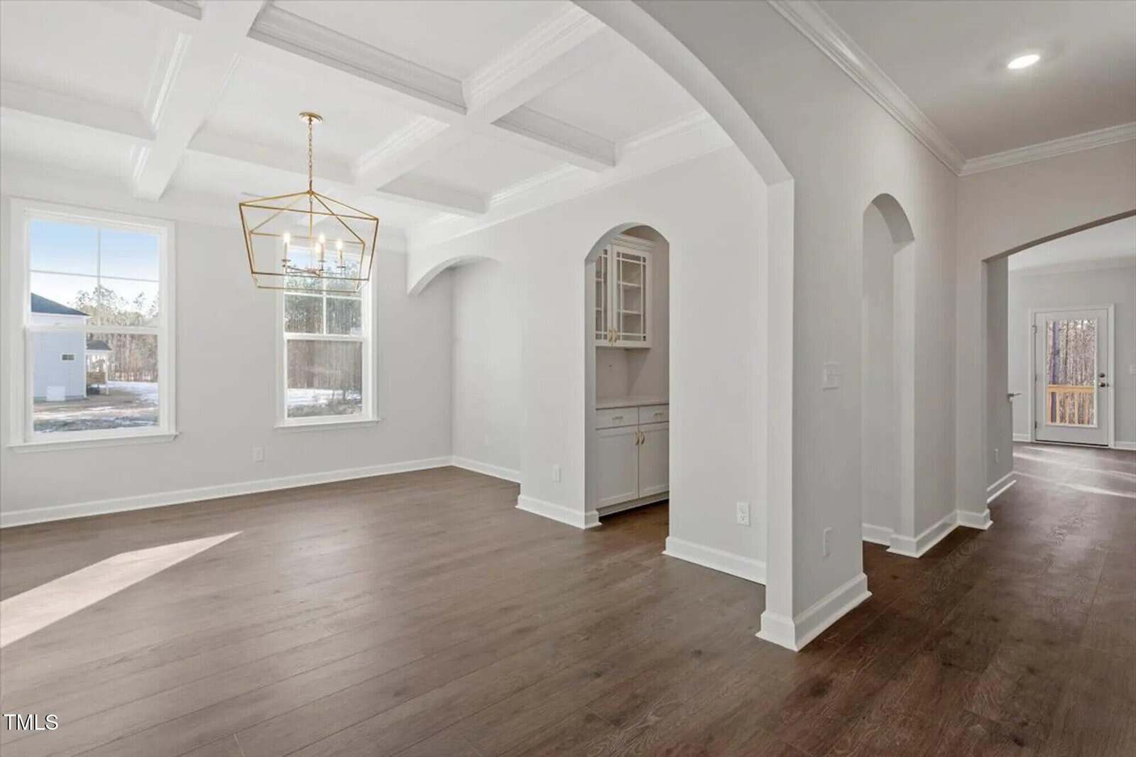 Bright, empty room with light gray walls, large windows, coffered ceiling, modern gold chandelier, and wood floors. Arched doorways lead to another room and a small built-in bar area with cabinetry.