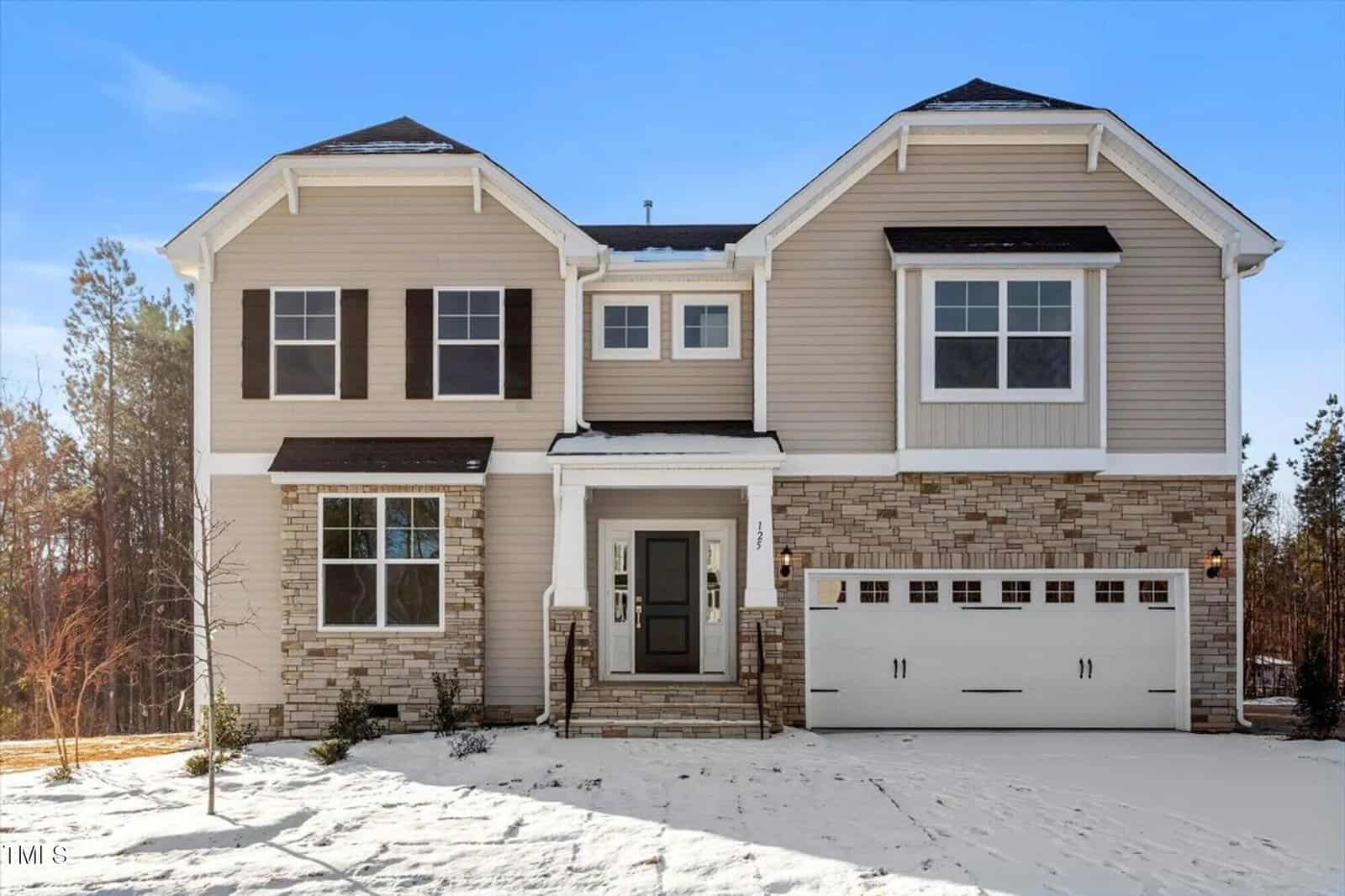 A two-story beige and white house with stone accents, double garage, black shutters, and snow covering the front yard and driveway on a clear day. Tall trees are visible in the background.