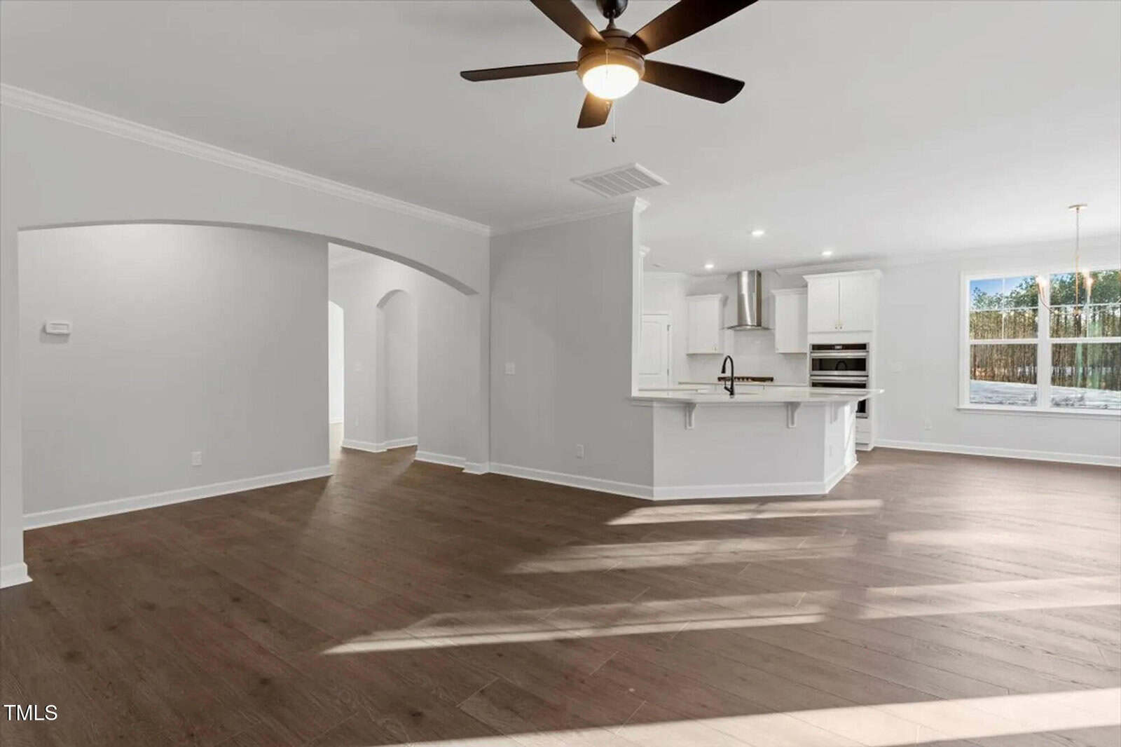 Spacious, empty room with wood flooring, white walls, ceiling fan, kitchen with white cabinets and stainless steel appliances, and large windows letting in natural light.