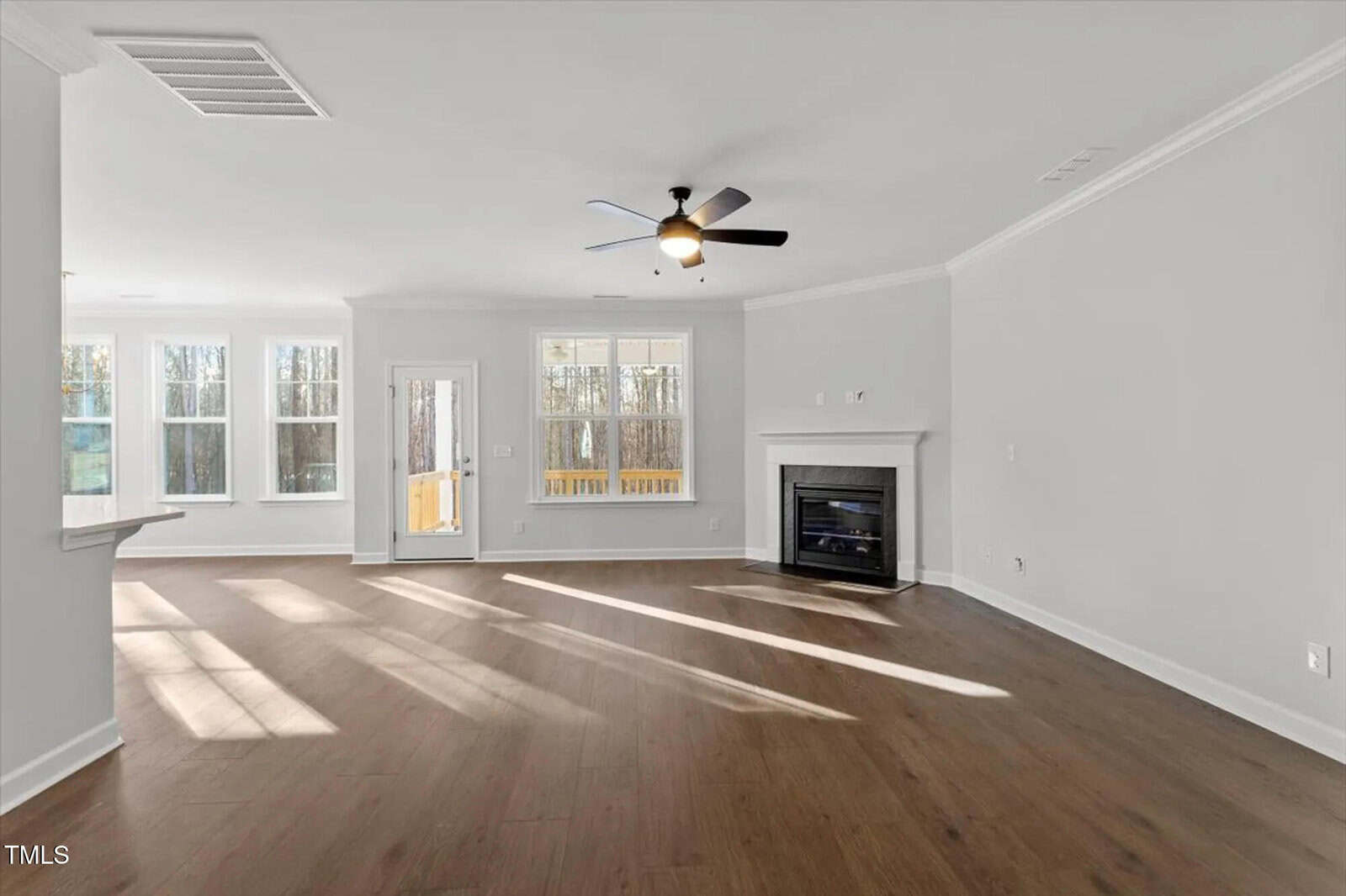 Bright, spacious living room with large windows, wood flooring, a ceiling fan, a corner fireplace, and white walls. Sunlight streams in, highlighting the open layout and view of a wooded area through the windows.