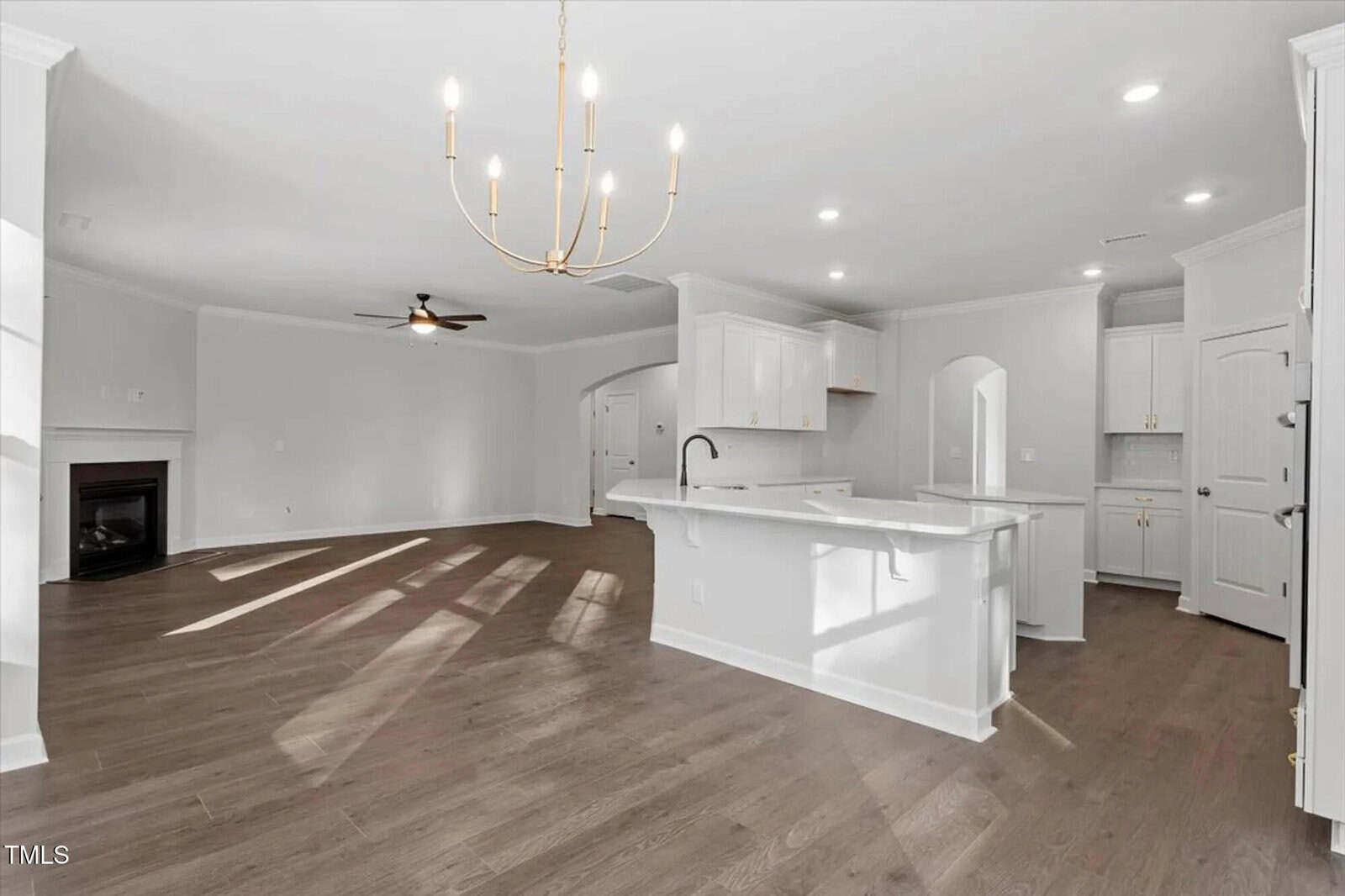 Open-concept kitchen and living room with white cabinets, a center island, wood floors, a chandelier, ceiling fan, fireplace, and large windows casting sunlight on the floor. Walls and ceiling are light-colored.
