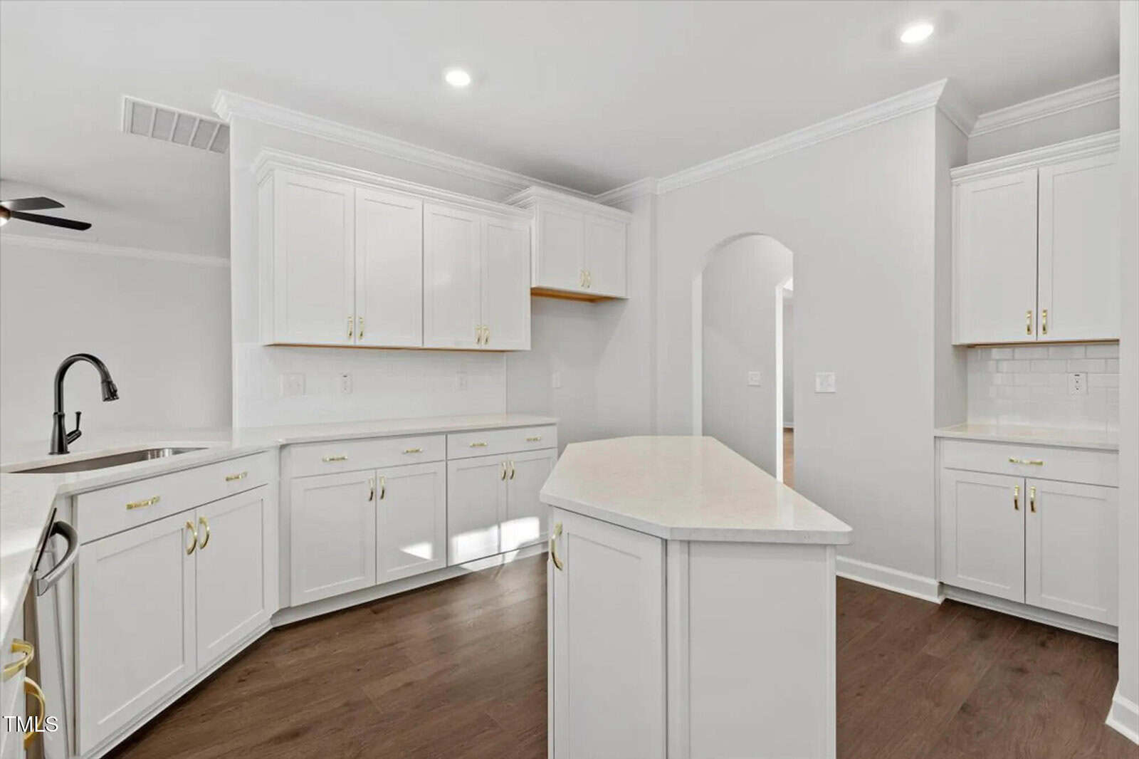 Bright, modern kitchen with white cabinets and gold handles, white countertops, an island in the center, stainless steel faucet, wood flooring, and an arched doorway leading to another room.
