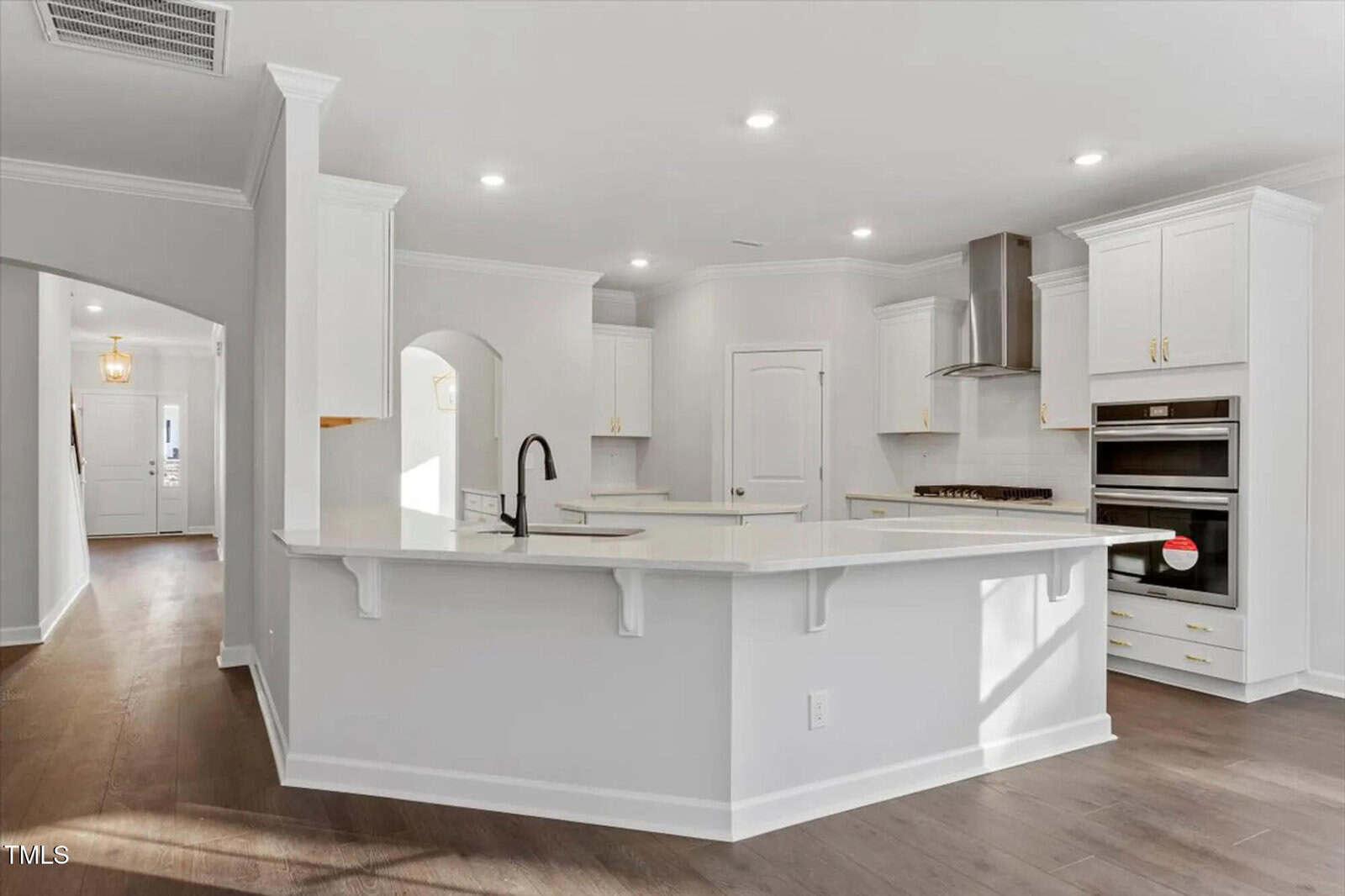 Modern kitchen with white cabinets, stainless steel appliances, a double oven, a large center island with a sink, and wood flooring. The space is bright with recessed lighting and an open layout.