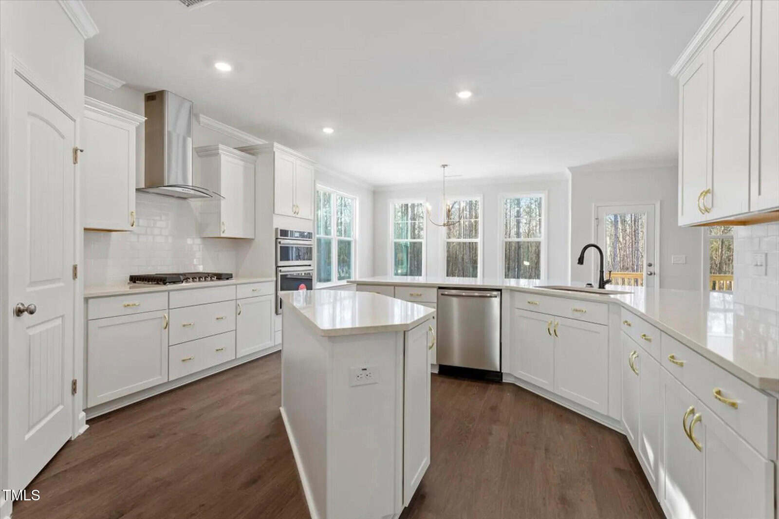 Spacious modern kitchen with white cabinets, gold handles, stainless steel appliances, center island, and large windows letting in natural light. Brown wood flooring contrasts with the bright, clean decor.