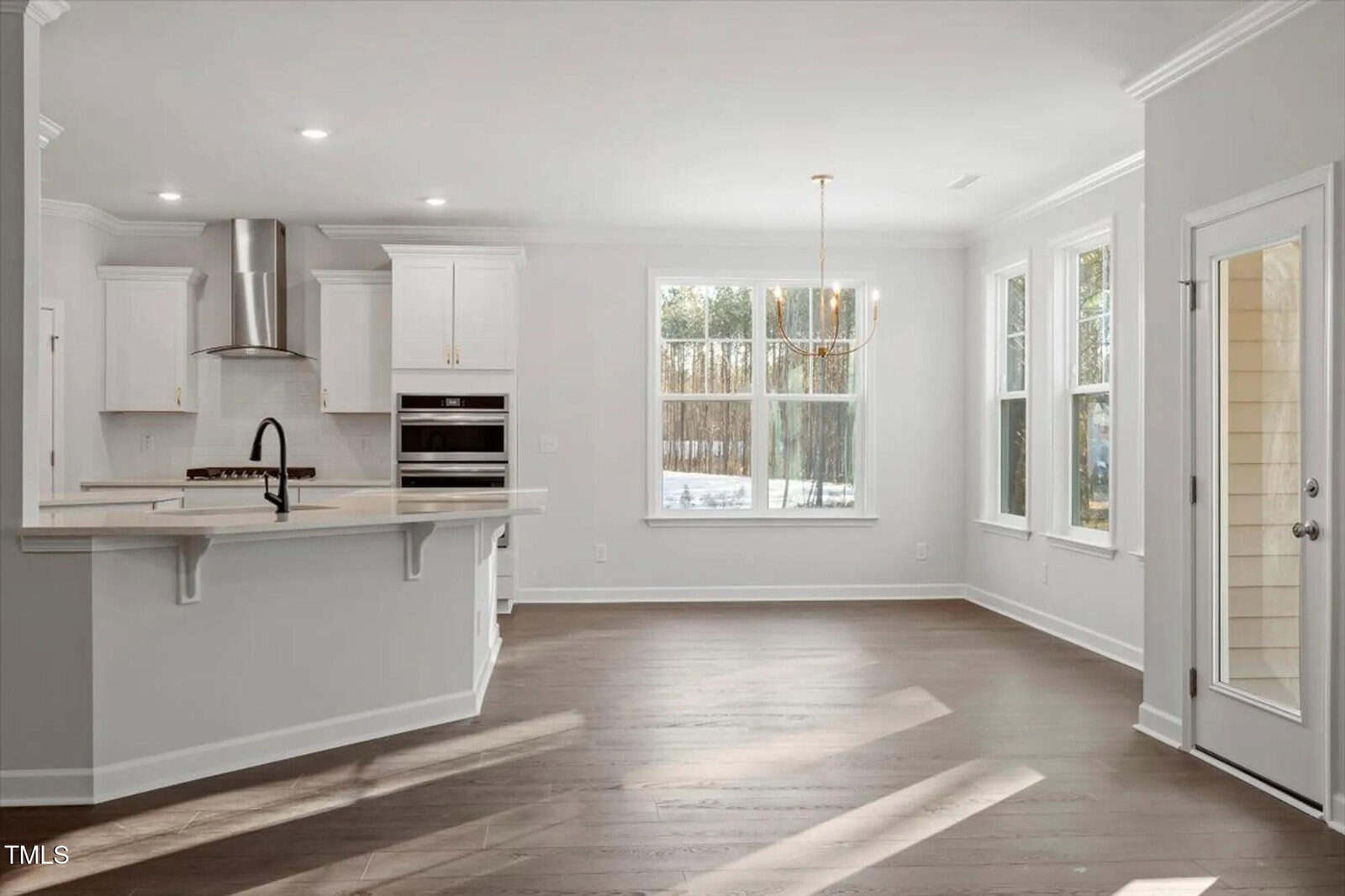 A modern, bright kitchen and dining area with white cabinets, stainless steel appliances, a large island with a dark faucet, pendant lighting, and large windows letting in natural light onto wood floors.