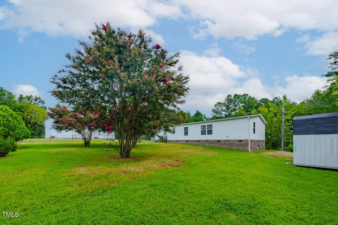 A white mobile home with brick foundation sits in a grassy yard. A large tree with pink flowers is beside it, and there is a small white shed in the right foreground. The sky is partly cloudy.